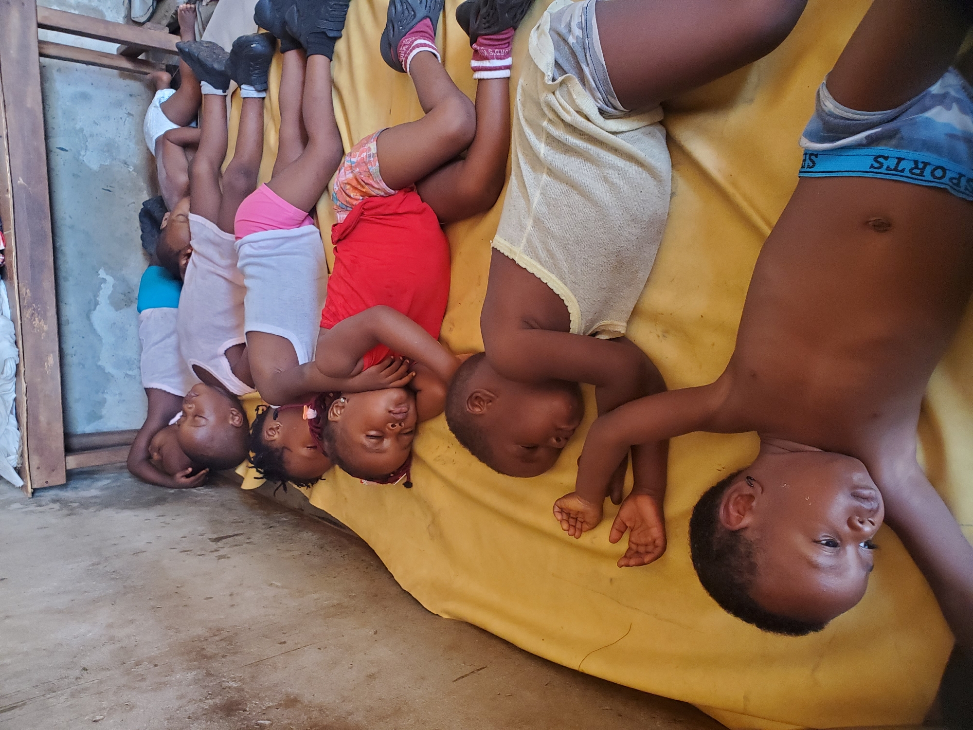In Ukwu-Abwa, Nigeria, a group of six children are lying head-to-toe on a yellow sheet on a concrete floor. They are arranged in a line, with their heads close together. Most appear to be sleeping, with their eyes closed. The children are dressed in various casual clothes, including shorts and T-shirts. Some of the children are wearing shoes. The room is sparsely furnished with a wooden frame visible in the background. The environment appears modest and simple.