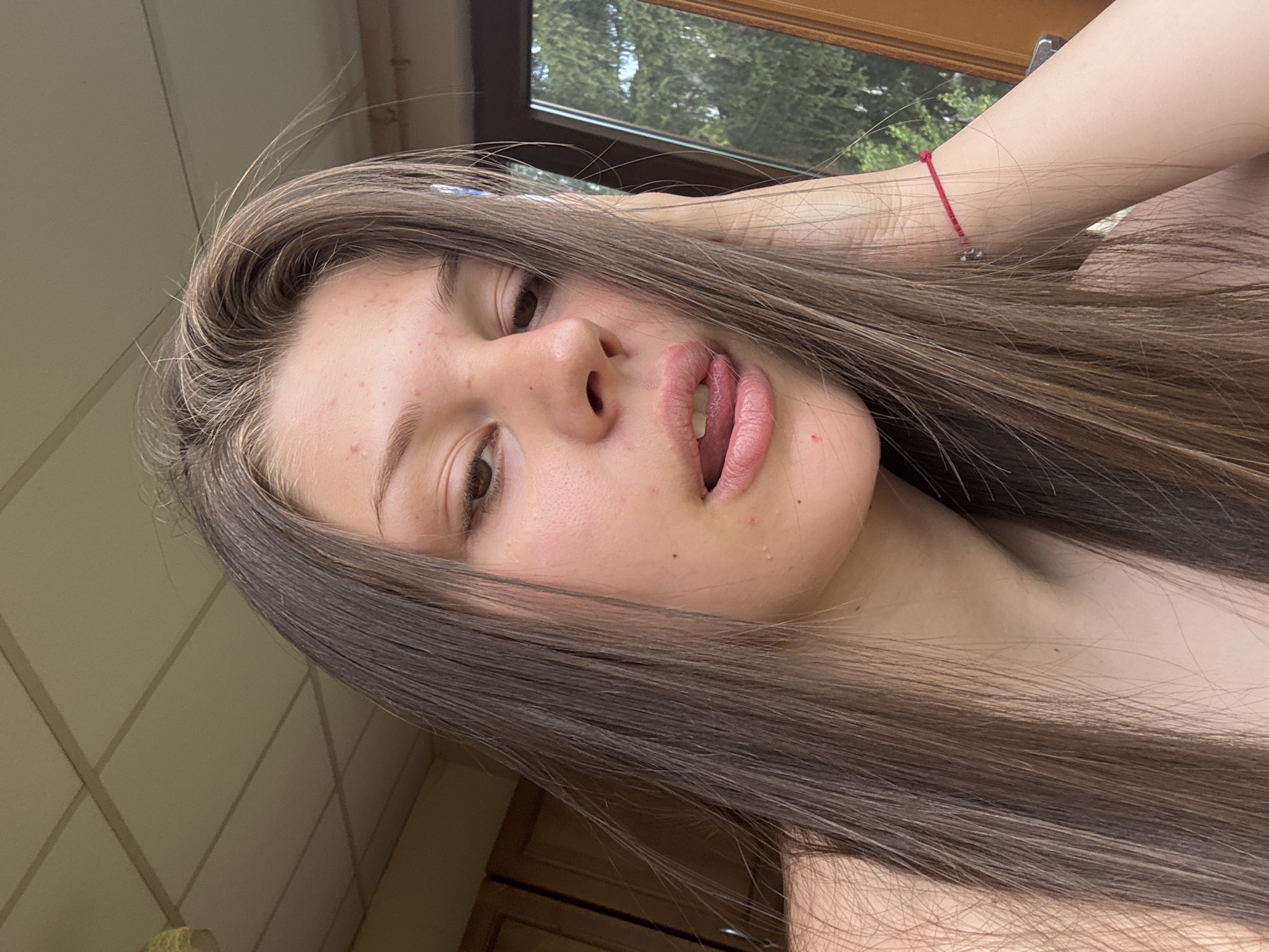 A young woman with long, dark brown hair is pictured in a close-up, tilted shot. Her hair frames her face, with some strands falling across her forehead and cheek. She has light skin with some visible blemishes and freckles. Her eyes are brown, and her lips are full and painted a soft pink. Her mouth is slightly open, revealing a glimpse of her tongue. A thin red bracelet with a small charm is visible on her wrist, peeking out from under her hair.
The background is out of focus, but appears to be an indoor setting with large windows showing greenery outside, suggesting a daytime scene. There are hints of light-colored walls with a diamond-patterned texture and a wooden frame, possibly a window or door. The lighting suggests natural light is present. The overall mood of the image is casual and relaxed, with the woman looking directly at the camera. No visible text is present in the image.