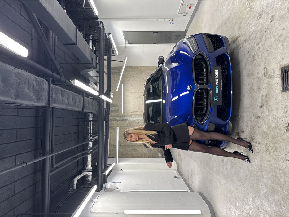 The image depicts an indoor scene within a luxury car showroom or detailing bay identified as Tiffany Motors in Warsaw, Poland. A single young adult blonde female stands to the left of a metallic blue BMW M8 coupe, which is parked centrally and facing the camera. The BMW's front license plate area explicitly displays "TIFFANY MOTORS." The female is dressed in a black blazer-style top worn as a dress, black sheer stockings with visible back seams, and black high-heeled pointed-toe pumps. Her left hand rests on her right forearm, while her right hand is slightly extended forward, and she maintains direct eye contact with the camera. The environment features a polished concrete floor with a visible drain grate, white textured side walls, a rough concrete rear wall, and a black industrial ceiling with exposed pipes, ventilation ducts, and numerous horizontal and angled white fluorescent strip lights providing bright, uniform illumination. No other individuals or direct interactions are depicted.