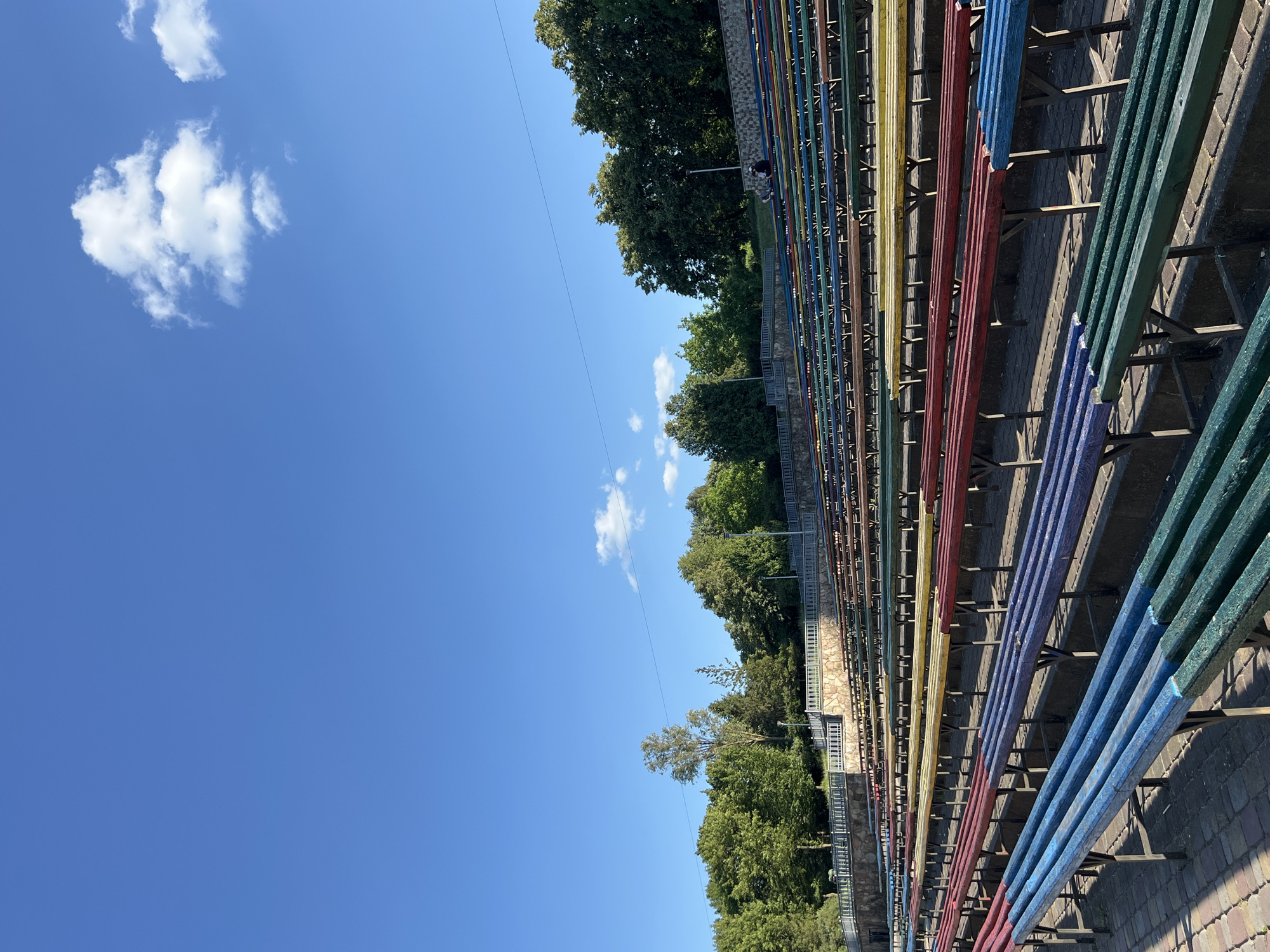 The image depicts an outdoor scene with a clear blue sky and scattered white clouds. In the foreground, a long row of brightly colored benches, painted in red, yellow, blue, and green, are arranged in tiered seating. The benches are constructed of wood and supported by metal frames. Behind the benches, there is a stone wall with a metal railing, and beyond that, lush green trees and foliage. A thin electrical wire stretches across the sky. The image is shot from a low angle, looking upwards towards the sky and the trees. The lighting suggests it is daytime, likely sunny with some shadows cast by the benches. The overall impression is of a public seating area, possibly in a park or stadium, in a green, outdoor setting. Two people are visible sitting on the benches, appearing to be relaxed. No text is visible in the image.