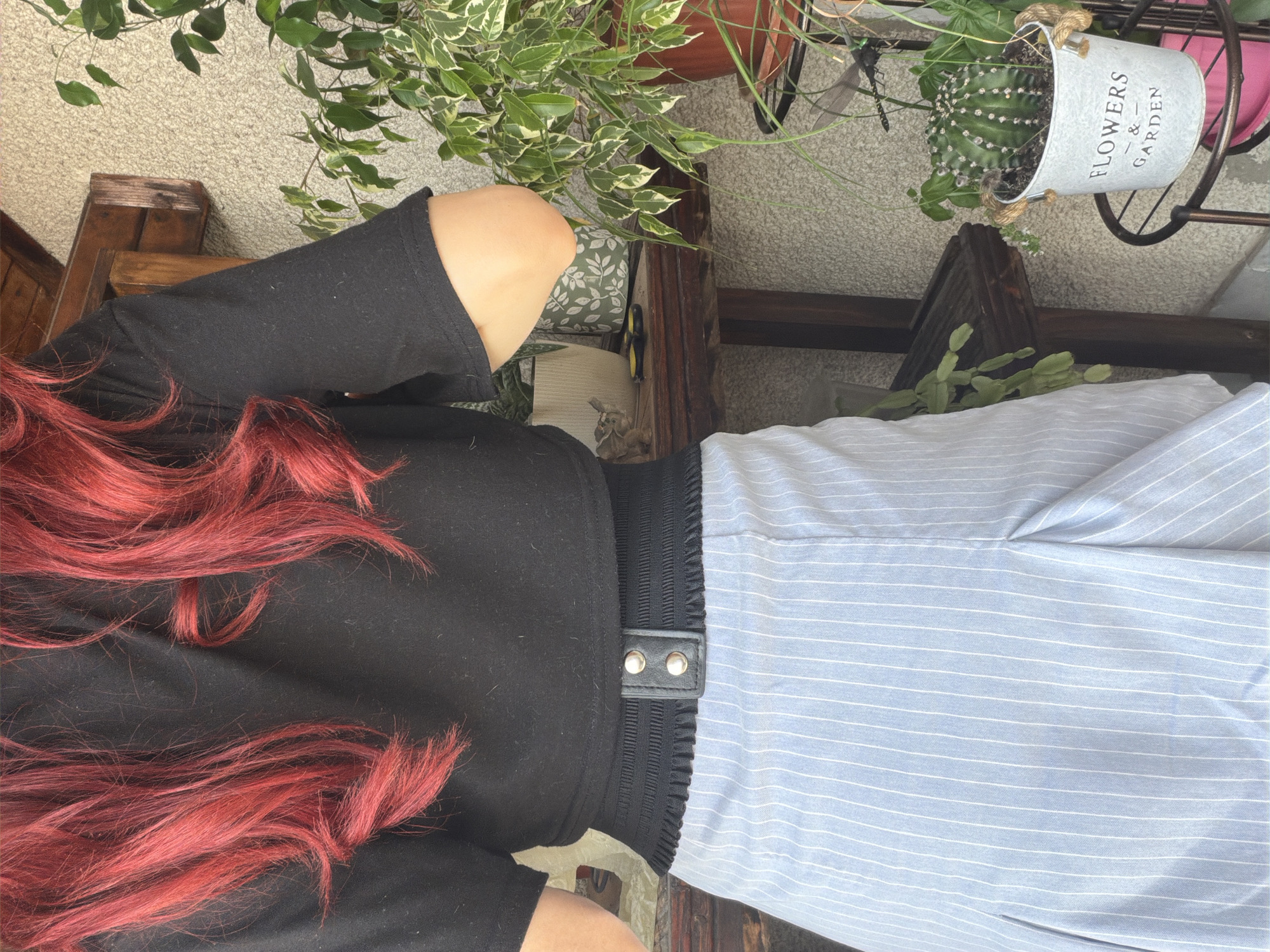 The image depicts the back of a person with vibrant red hair, wearing a black crop top and a light blue pinstripe skirt. The skirt features pleats and a black waistband with a decorative buckle. The person is standing outdoors, near a textured wall, with a wooden shelf holding various potted plants. A cactus in a bucket-like planter with "FLOWERS & GARDEN" inscribed on it is visible on a tiered stand. The setting appears to be a residential balcony or patio. The exact location in Svilengrad, Bulgaria, is not identifiable from the visual information provided.