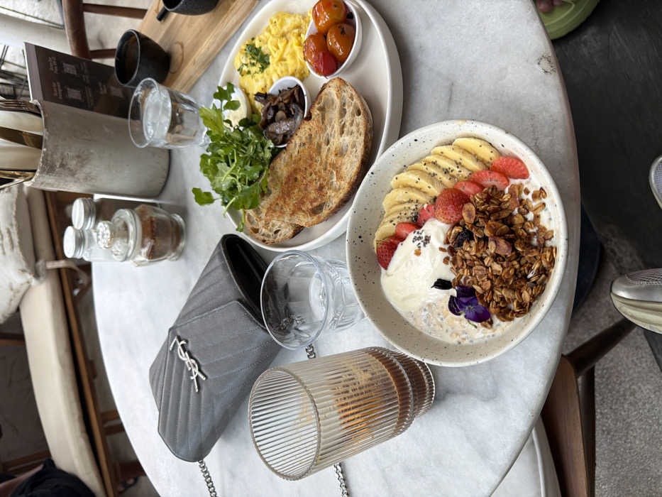 A marble table displays two plates of food: one with scrambled eggs, toast, and mushrooms, the other a bowl of yogurt, granola, strawberries, and bananas. Next to the food are two glasses of water, a ridged glass container, and a gray YSL purse with a chain strap. Condiment jars, a menu, and silverware are also visible on the table. Part of a chair and a person’s foot are in the background.