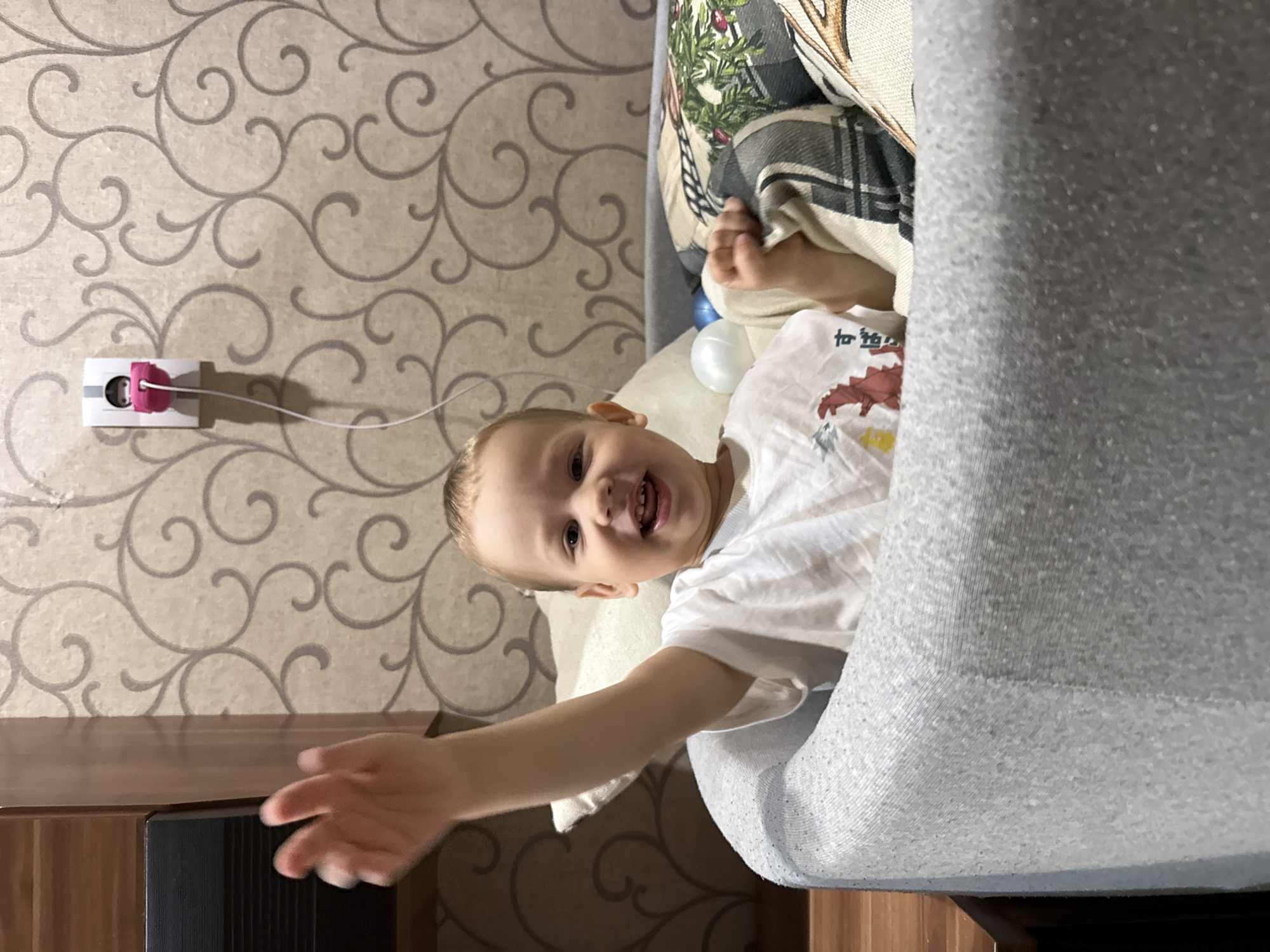 A young child lies sideways on a grey sofa in a room. The child is wearing a white t-shirt with a dinosaur print. The child smiles with mouth open and left arm outstretched. The background includes beige patterned wallpaper, a white electrical outlet with a pink cord organizer, and a wooden cabinet. A patterned blanket is partially visible on the sofa. Location is Rusanovo, Russia, based on provided coordinates.