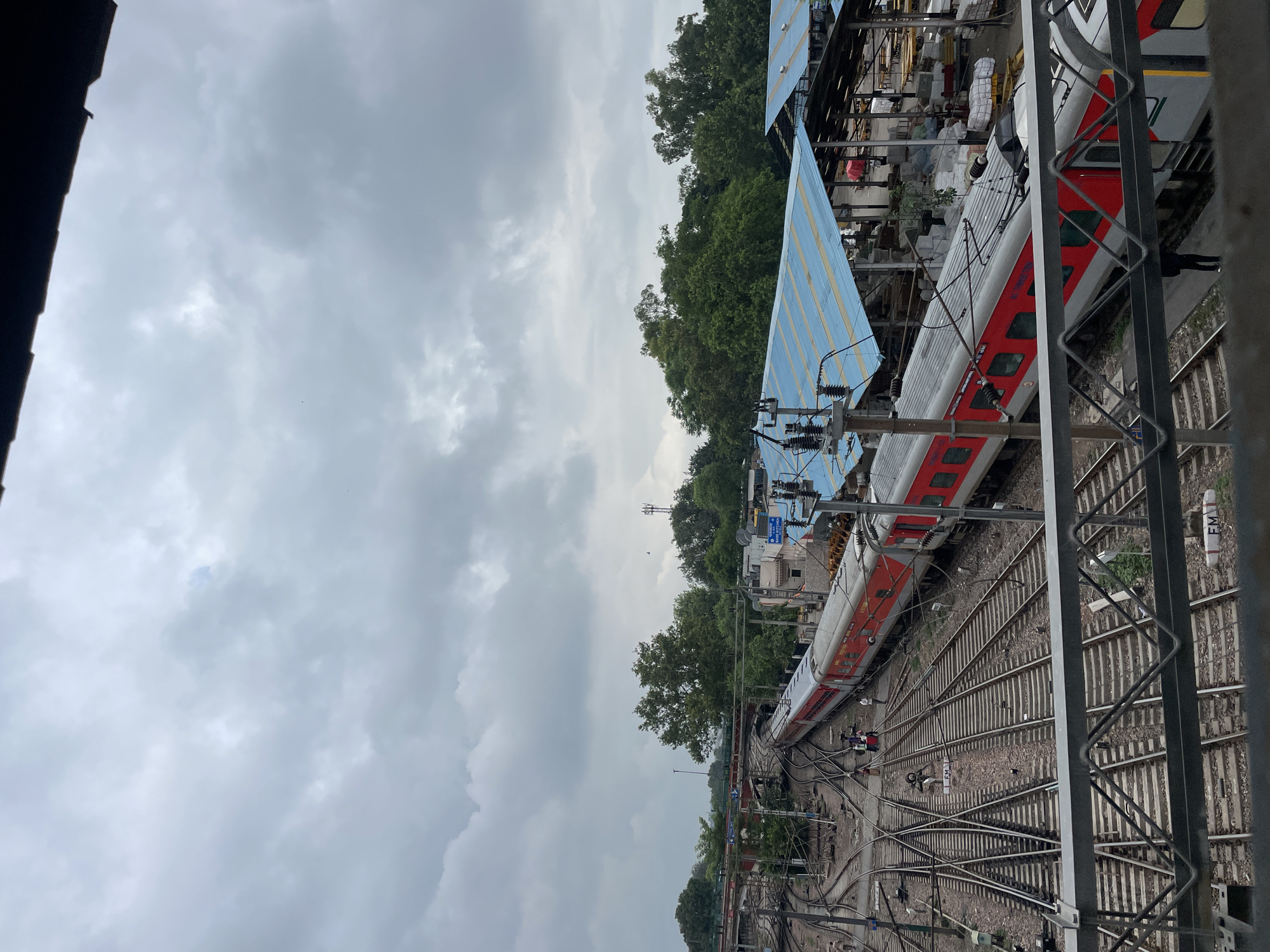 The image depicts a train station in New Delhi, India, under a cloudy sky. A red and silver train is positioned on multiple tracks, with a complex network of railway lines extending into the distance. In the background, there are buildings with blue roofs, trees, and a telecommunication tower. Several people are visible on the platform and near the train. The foreground features a metal railing of a bridge or walkway overlooking the train tracks. The scene suggests an active railway hub in an urban environment.