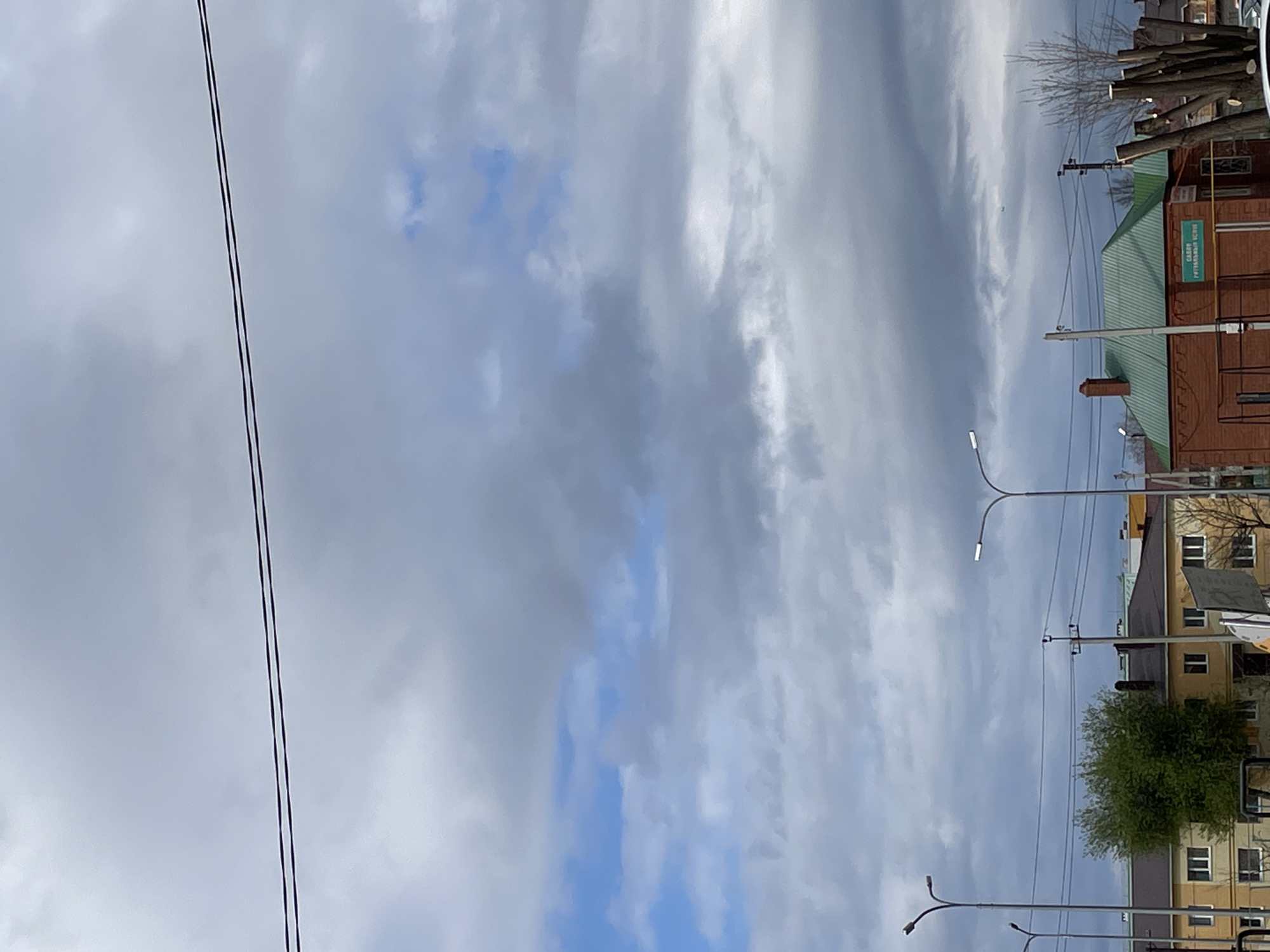 The image shows a street in Urussu, Russia. Overhead power lines stretch across the left side of the frame. On the right side of the frame are buildings, streetlights, and trees. The sky is overcast with a mix of gray and blue clouds.