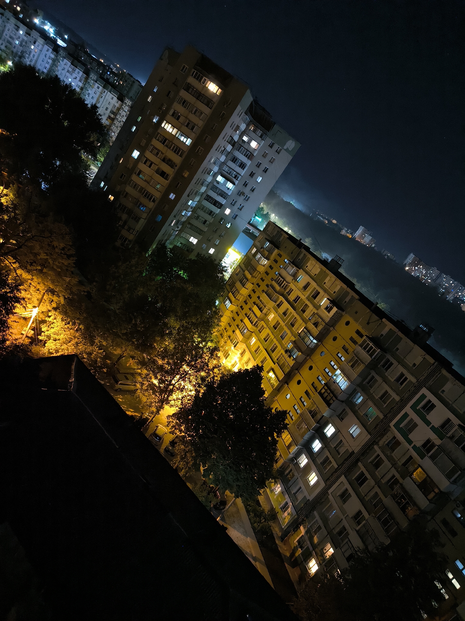 The image is an aerial, nighttime view of a residential area in Chișinău, Moldova. The perspective is tilted, looking down and across a collection of multi-story apartment buildings. The buildings are illuminated by the lights from their windows, creating a warm glow against the dark sky. Some streetlights and the lights from distant buildings are also visible, suggesting a moderately urban environment. Trees, some with yellow light cast upon them, are interspersed between the buildings, indicating green spaces within the city. The overall impression is one of a quiet, lit-up city at night. There are no people or specific actions depicted.