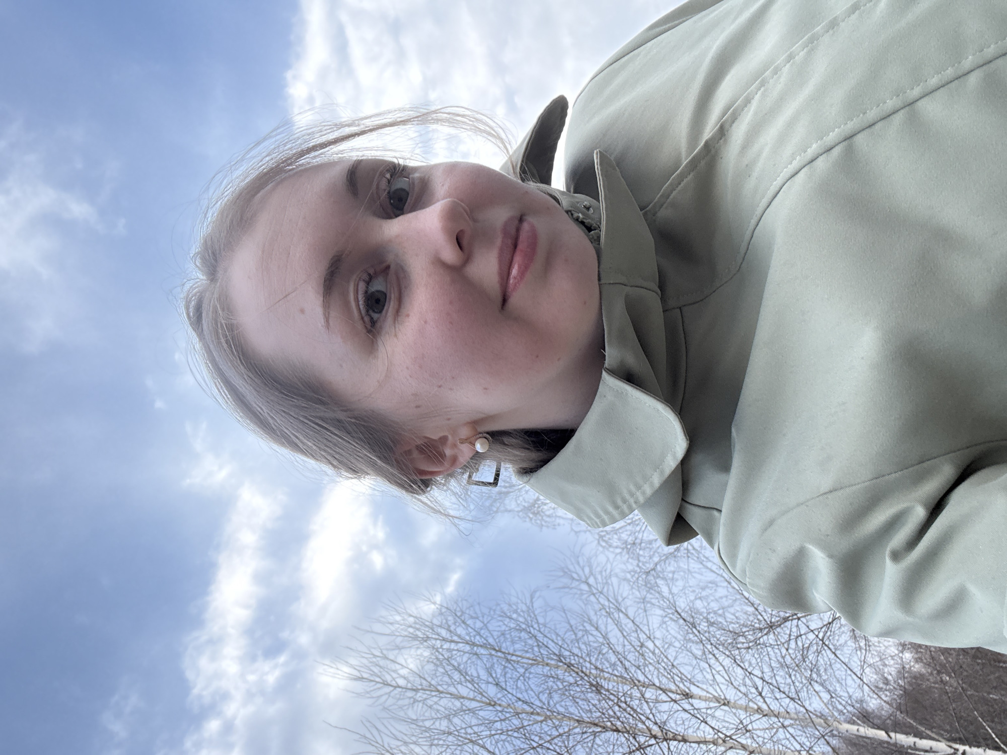 A selfie-style photograph captures a young woman with light-colored hair and fair skin, featuring light freckles across her cheeks. She has light eyes and her lips are slightly parted, giving her a neutral to slightly expressive look as she gazes directly into the camera. She wears a light sage green or pale olive-colored collared jacket. On her visible left ear, she has a small pearl stud earring and a larger, square-shaped dangle earring.
The image is taken from a low angle, looking upwards, and is oriented sideways, with her head on the left side of the frame and her body extending towards the right. The background is dominated by a bright blue sky with scattered white clouds, indicating clear daytime weather. In the lower part of the frame, particularly towards the right, the bare branches of deciduous trees are visible against the sky, suggesting a colder season like late autumn, winter, or early spring. The setting appears to be an outdoor, natural environment. No text is visible in the image.