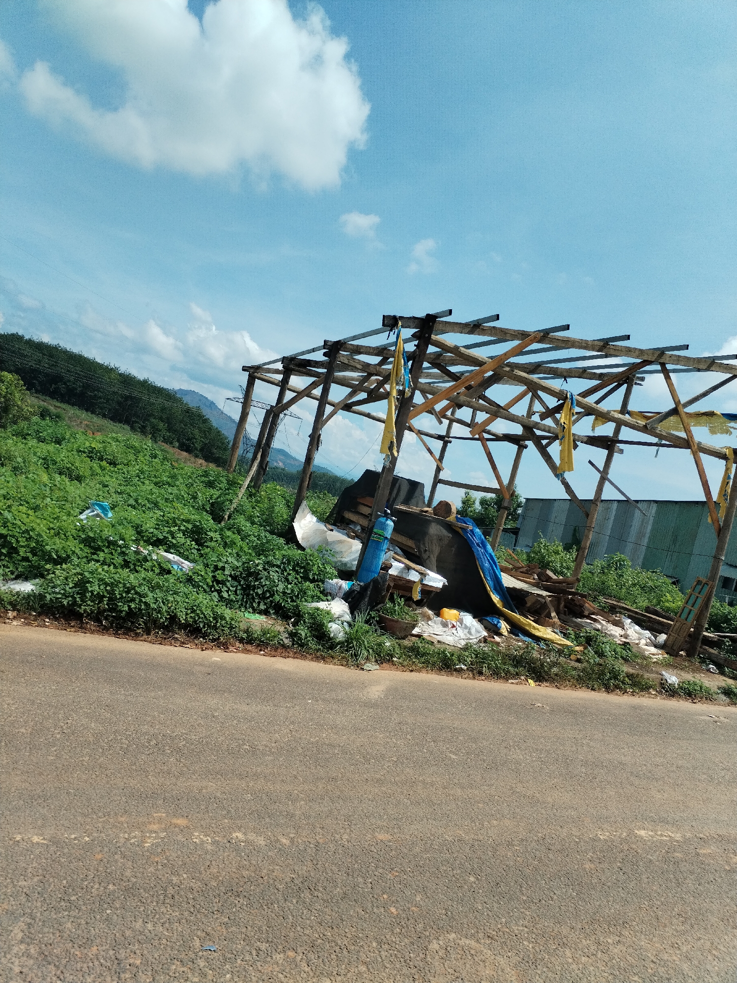 The image shows a scene in Bờ Y, Vietnam. A dilapidated wooden structure with a partially collapsed roof stands adjacent to a paved road. Debris, including blue and yellow tarps, scattered pieces of wood, and various containers, are piled within and around the structure. Dense green vegetation covers the nearby hillside. In the background, a corrugated metal building is visible, as well as power lines. The sky is blue with scattered white clouds.