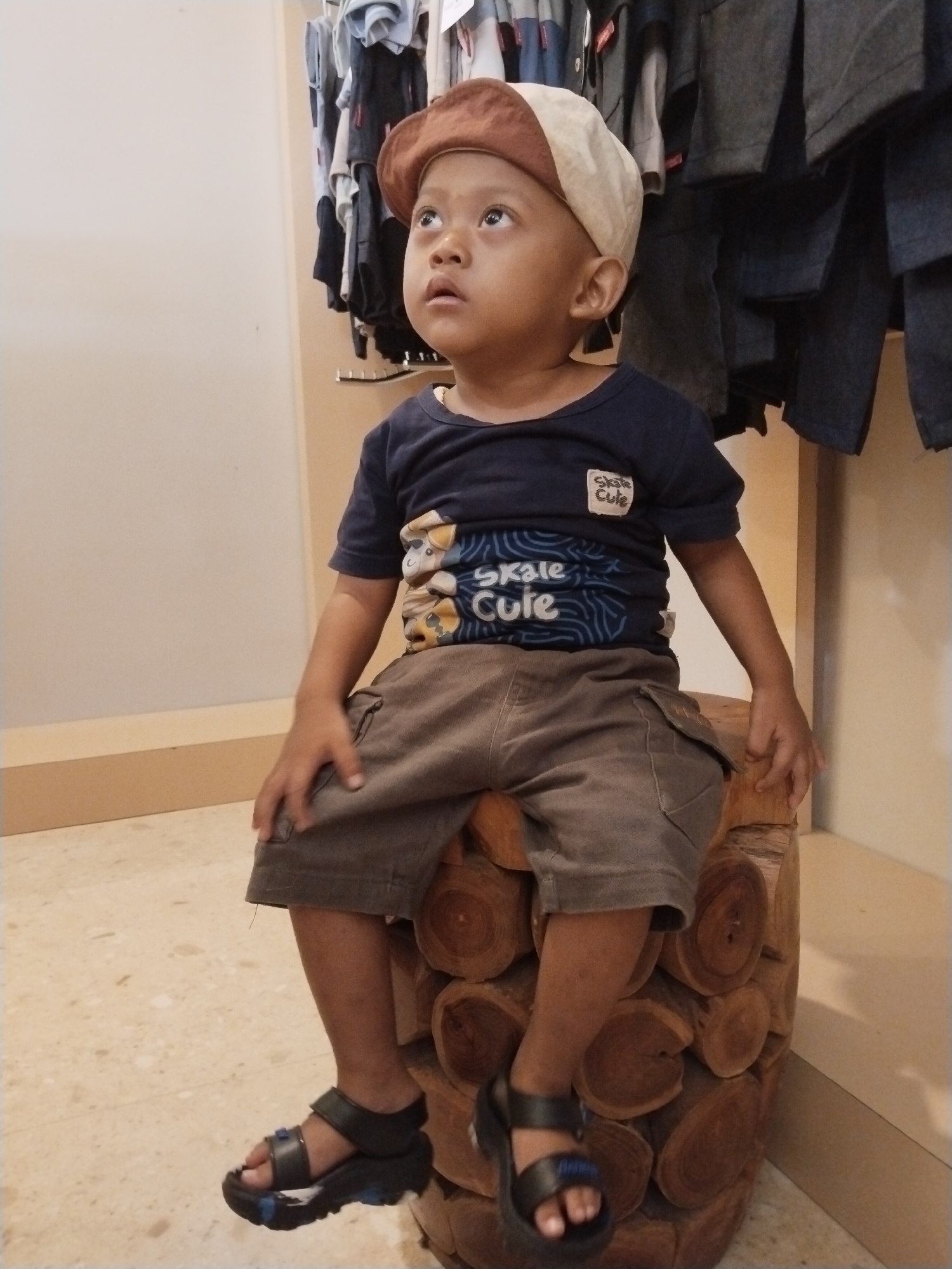A young child with Down syndrome, wearing a brown and cream colored flat cap, a dark blue t-shirt with graphics of cartoon characters and the text "Skale Cute", and brown shorts, sits on a rustic, cylindrical wooden stool made of stacked logs. The child is looking upwards and to the left. The background features clothing on racks, suggesting a retail environment, possibly in Kecamatan Pati, Indonesia. The floor is tiled with a light beige pattern. The lighting is ambient, indicating an indoor setting.