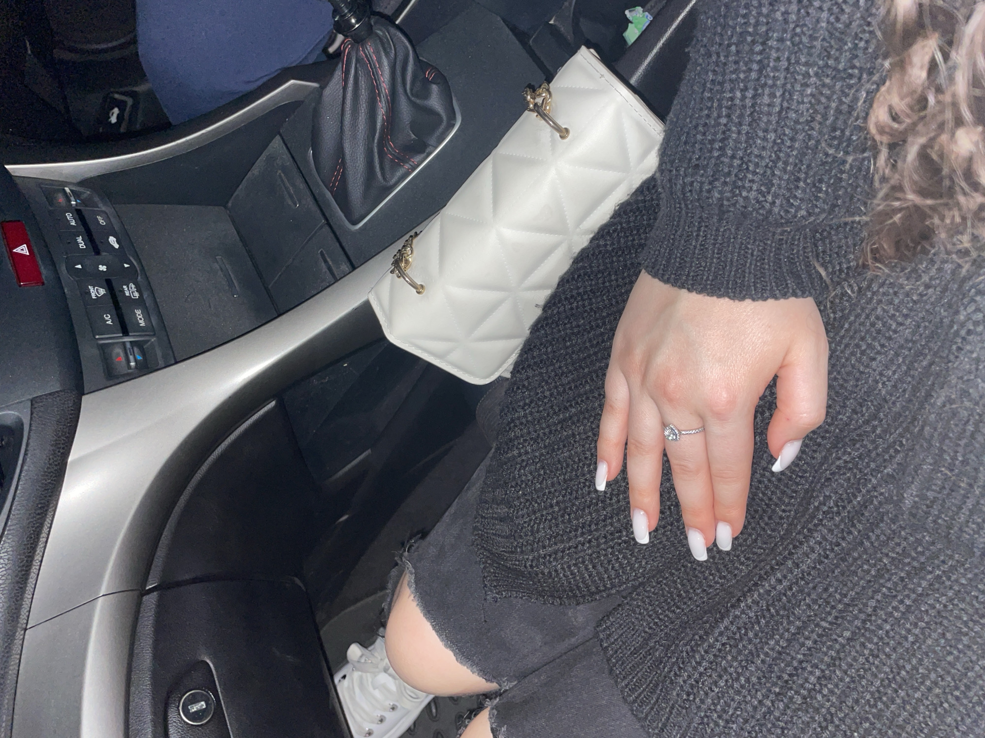 A partial view of a car interior showing a person's arm and hand. The person is wearing a dark gray knit sweater. The hand is resting on the sweater-clad arm and features white-tipped manicured nails and a silver ring with a clear stone. A white quilted purse with gold hardware is positioned near the car's gearshift. The car's interior includes elements like the gearshift knob with red stitching, climate control panel, and dashboard. A glimpse of the person's distressed black shorts and a white sneaker can be seen near the knee. The photo does not reveal any elements to identify the location as Zlatograd, Bulgaria.