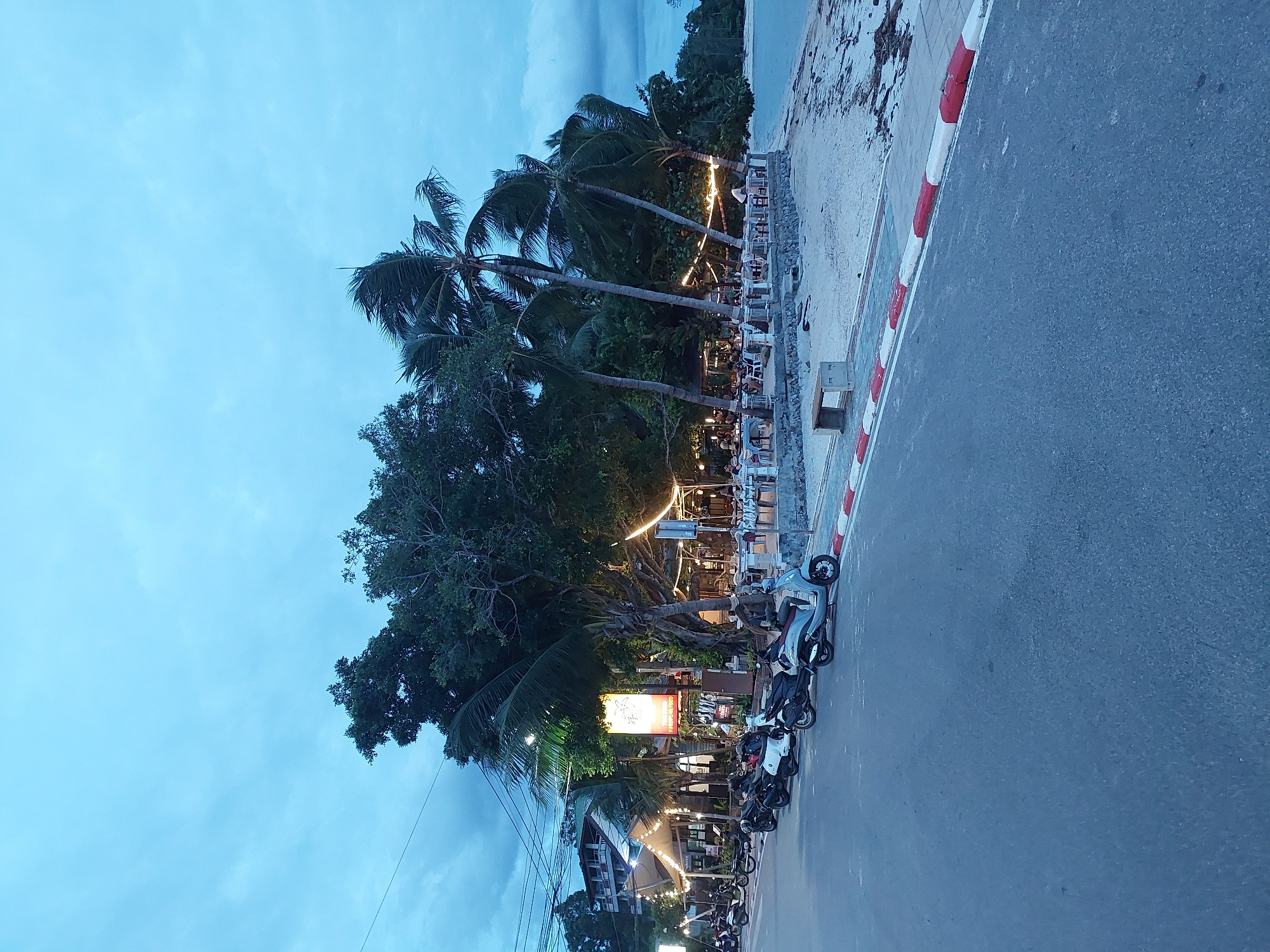 The image depicts a waterfront scene in the Ko Samui District of Thailand, likely during dusk or early evening. A paved road runs along the left side of the frame, lined with parked scooters. Beyond the road, a sandy beach area is visible, with an outdoor restaurant set up with tables and chairs. String lights illuminate the restaurant area, creating a warm ambiance. Palm trees are scattered throughout the scene, some leaning towards the water. In the background, a multi-story building can be seen. The sky is overcast, with hints of a fading sunset or approaching night. The overall impression is that of a relaxed, tropical evening by the sea.