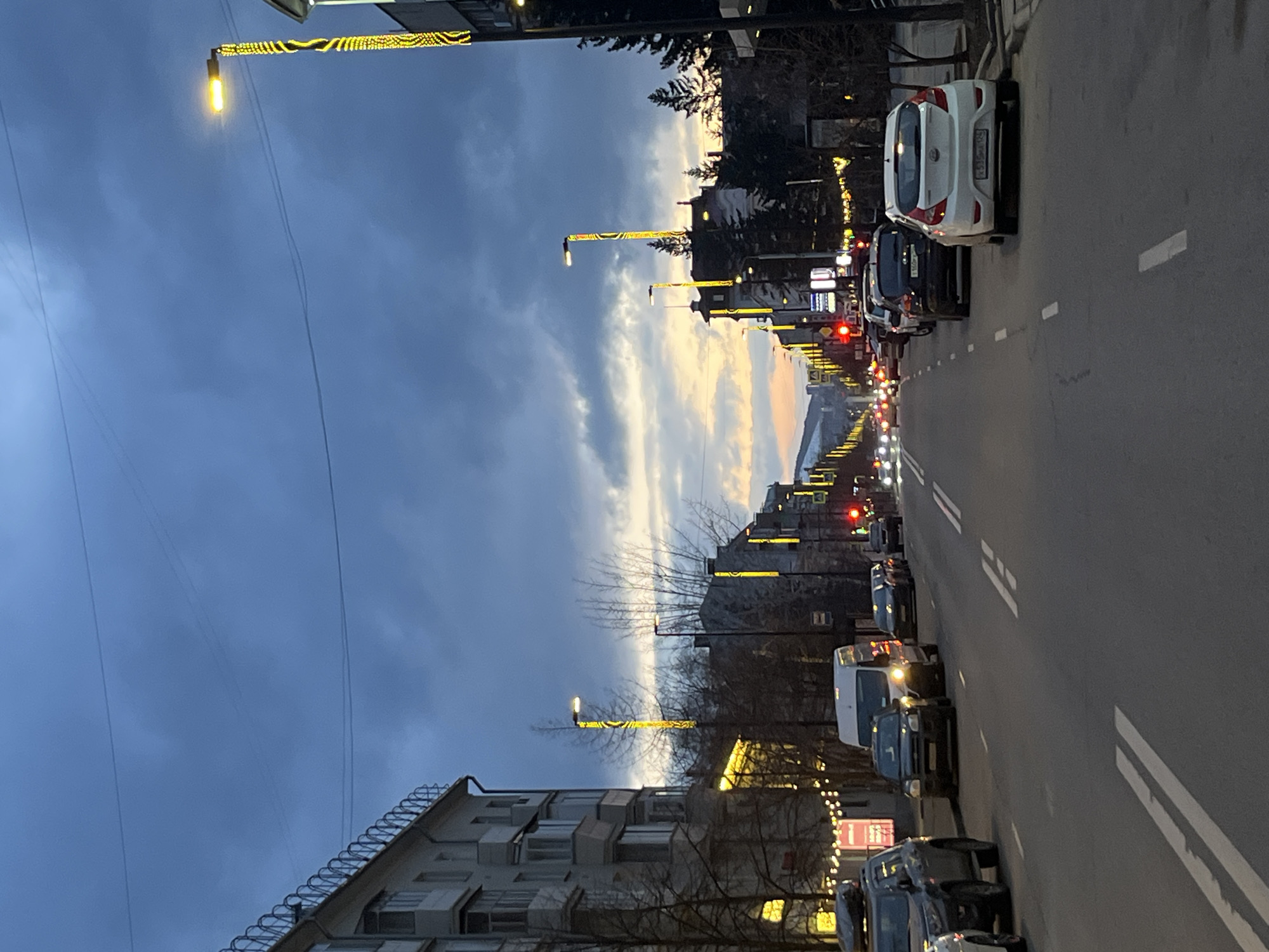 The image captures an urban street scene during dusk or early night in Krasnoyarsk, Russia. The sky, transitioning from deep blue to lighter, warmer tones on the horizon, is partly cloudy. Streetlights and decorative yellow lights lining the road are illuminated, casting a warm glow against the fading daylight.
The asphalt road, marked with white lines, extends into the distance. Several vehicles are visible; some are parked along the right side of the street, while others appear to be in traffic with their headlights and taillights on. A white hatchback, prominently parked on the right, displays a Russian license plate "O 519 MO 05". Other cars, including a dark SUV/minivan and a white van, are further down the road.
On the left side of the frame, a multi-story building with numerous windows and balconies is visible. Bare trees, likely indicating late autumn or winter, line the street between the buildings and the road. The overall impression is one of a calm, everyday city evening, possibly with a festive touch due to the decorative street lighting. No people are distinctly visible.