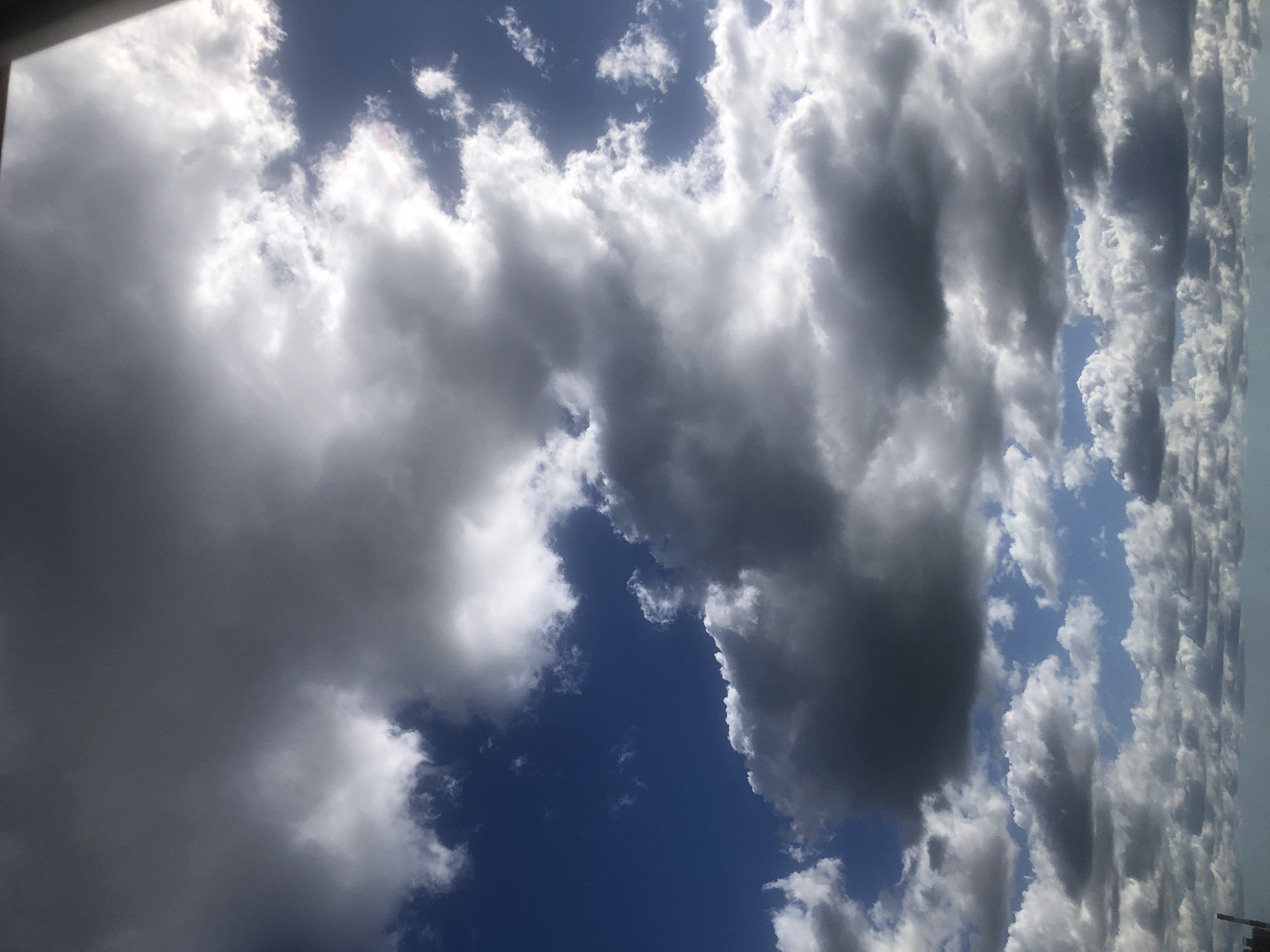 A low-angle shot captures a dynamic sky filled with a mix of dark gray and bright white clouds against a vivid blue sky. The clouds are dense and varied in texture, with some appearing puffy and illuminated by sunlight, while others are heavy and shadowed. The sunlight is bright and casts dramatic highlights on the cloud formations, particularly in the upper right portion of the image. The scene suggests an overcast but bright day, possibly with a storm approaching or passing. The lower edge of the image shows a faint hint of a distant, hazy horizon, possibly indicating a cityscape or landmass, but the focus is entirely on the sky. No people or specific landmarks are clearly visible.