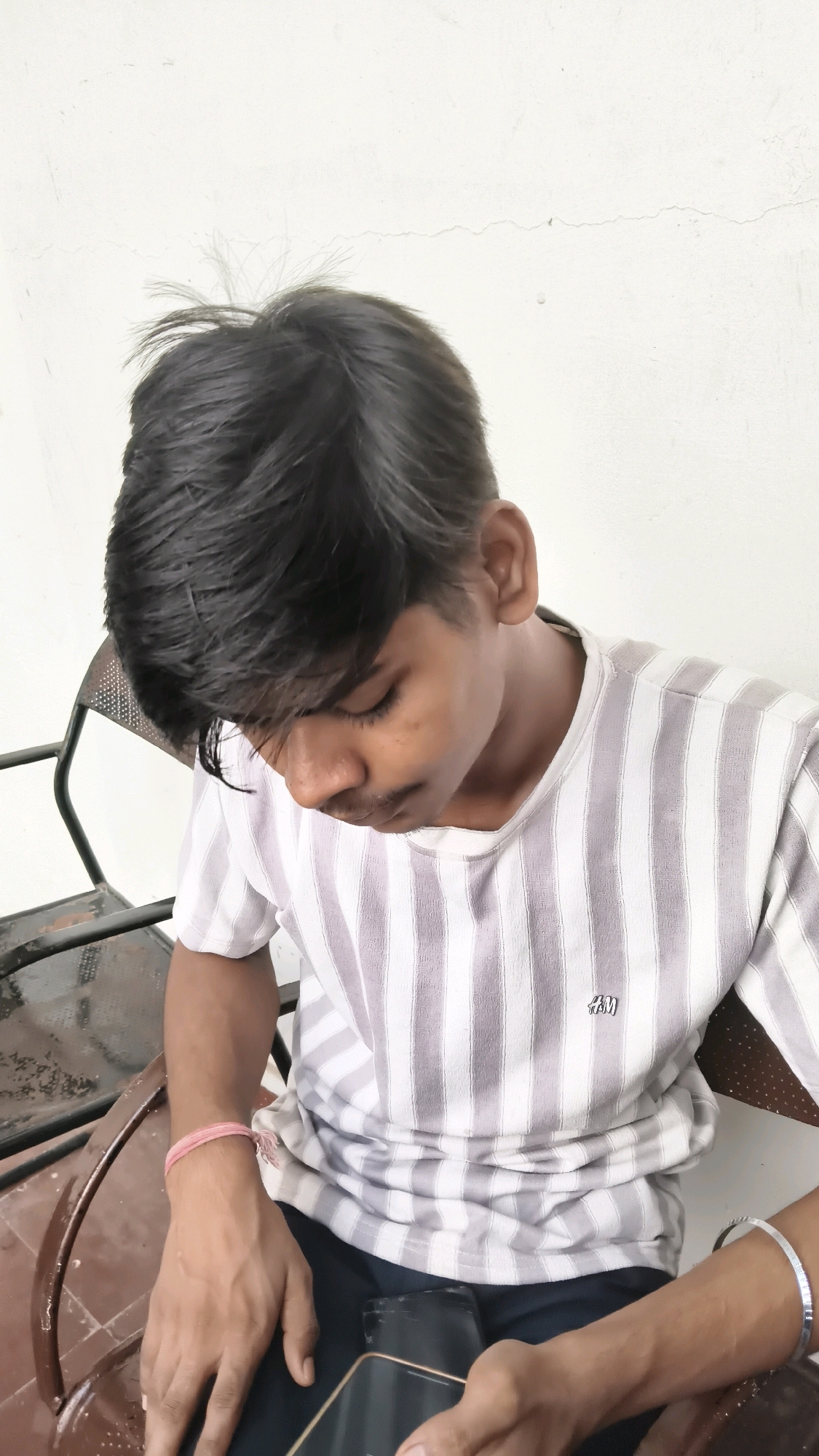 A young man with dark hair is sitting on a metal chair in front of a white wall. He is wearing a horizontally striped t-shirt and dark pants. His head is bowed, and he is looking down at a phone in his hands. A pink bracelet is visible on his left wrist. The background shows a part of a metal chair and a white wall with a crack. The scene appears to be outdoors, possibly in a city like Libra, India, given the context.