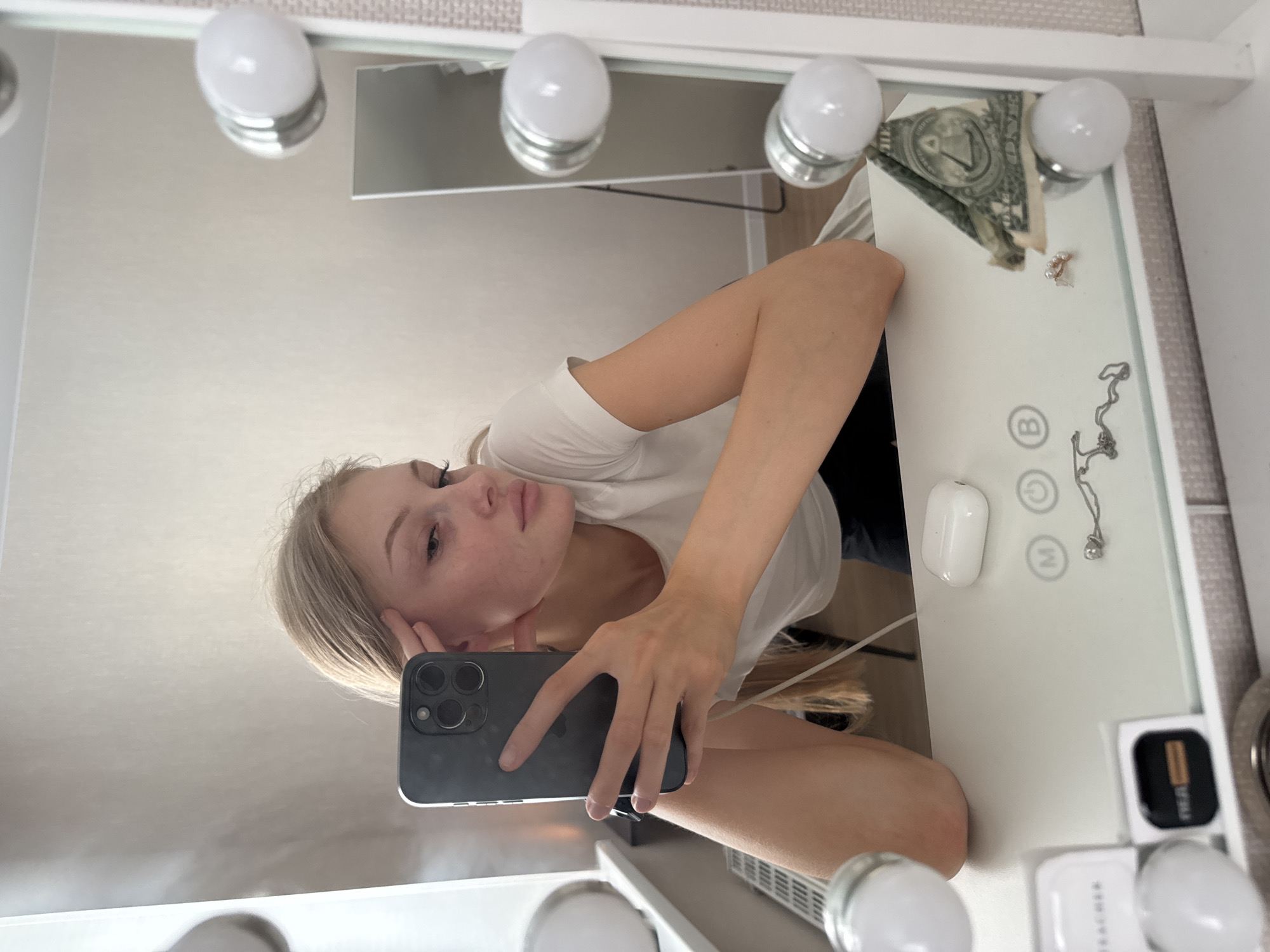 A young woman with blonde hair is taking a selfie in a mirror. She is leaning on a white vanity table and holding a smartphone in her left hand. The mirror is framed with lights, and a second, taller mirror is visible in the background. On the vanity table, there are several items, including a pair of wireless earbuds, a necklace, a ring, and a folded US dollar bill. The overall setting appears to be a dressing room or bedroom. There is no specific information in the image to suggest the location is in the city of Perm, Russia.