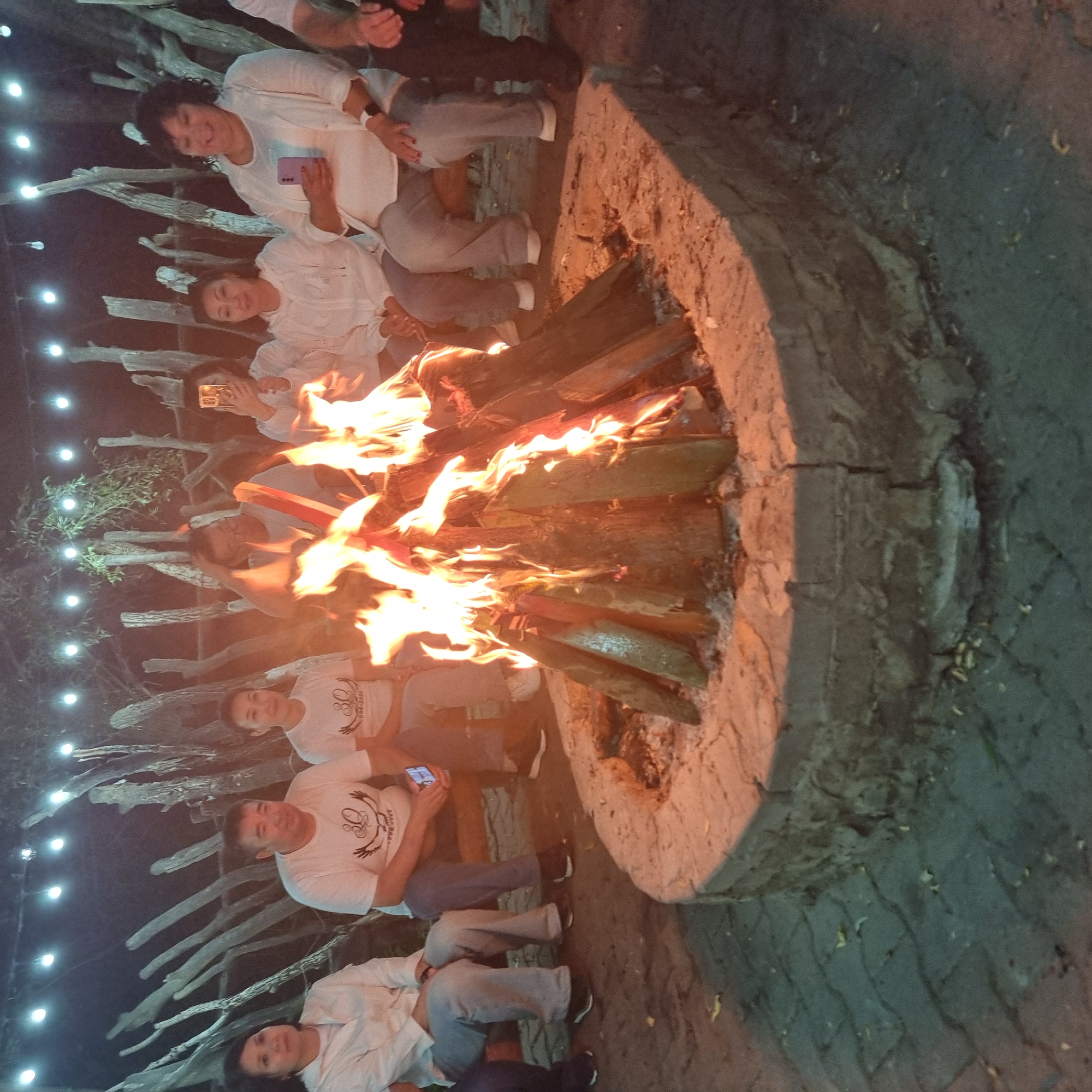 The image shows a group of eight people, a campfire, and an outdoor seating area. The people are sitting on a bench around a circular fire pit. The fire pit is made of stone and is filled with burning wood. The people are of various ages and ethnicities, and they are all dressed in casual clothing. The background of the image shows a rustic wooden fence and some trees. The lighting suggests it is evening or nighttime, with string lights visible in the background. A woman on the far right is holding a phone, and another woman in the middle is also holding a phone. Two men and six women are present. The people appear to be enjoying themselves, with some smiling and interacting with each other. It is not possible to determine the specific location in the city of Aktobe, Kazakhstan from the image alone.