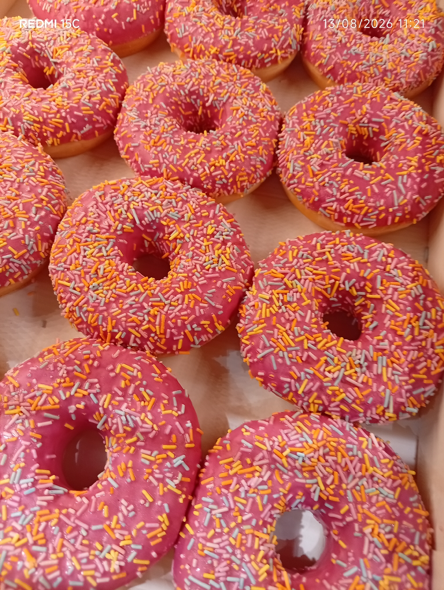 A close-up overhead shot shows a box of donuts with pink frosting and colorful sprinkles. The donuts are arranged in a staggered pattern. The lighting is bright, and there are no discernible background elements or people. The image is a still life of baked goods.