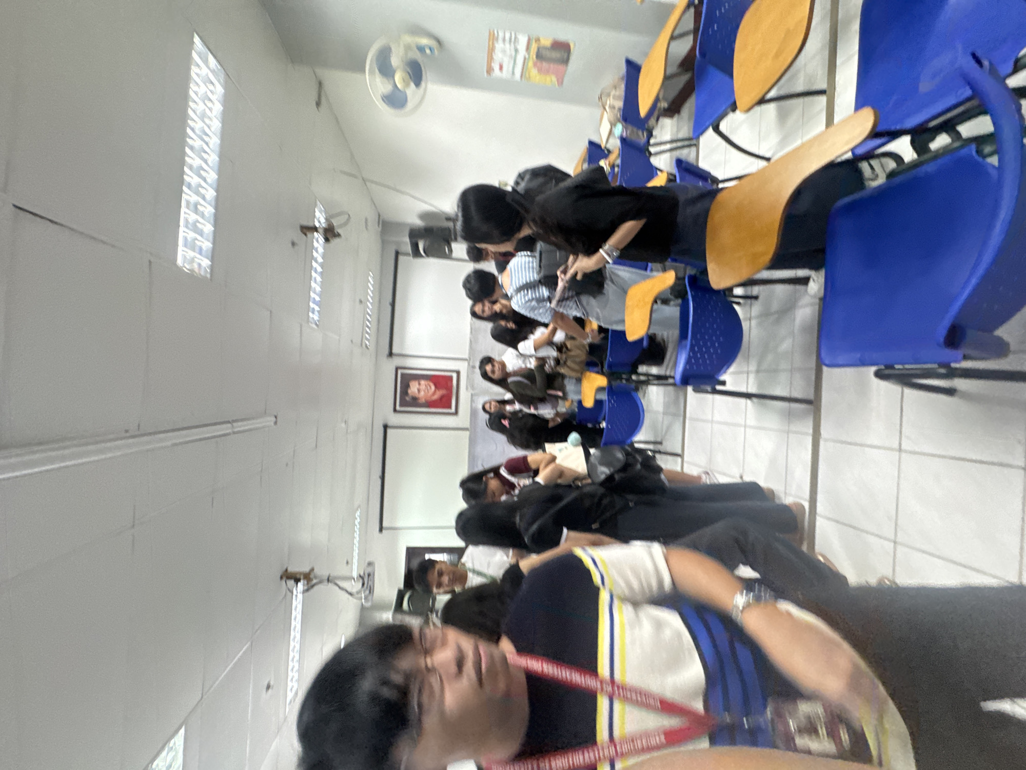 The image depicts a classroom setting in Davao City, Philippines. Rows of students are seated in blue and yellow chairs, facing a projector screen at the front of the room. A portrait hangs on the wall near the screen. A ceiling fan is visible. The fluorescent lighting fixtures are visible on the ceiling. The photograph appears to have been taken by a student in the foreground.