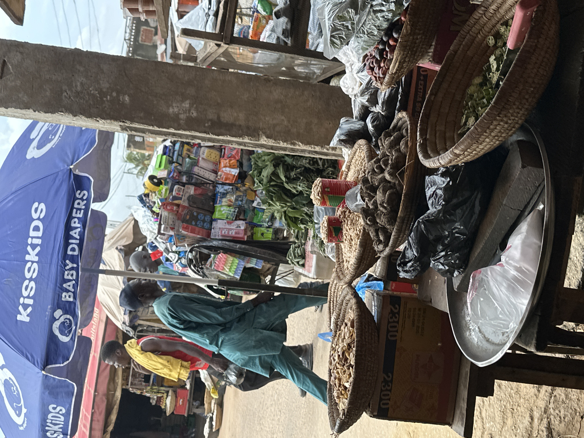 The image depicts a bustling market scene in Narayi, Nigeria, under a large blue umbrella advertising "KissKids Baby Diapers." In the foreground, several woven baskets overflow with goods. One basket contains a pile of pale, dry seeds or nuts, another holds dark, clumpy root vegetables, and a third is filled with chopped green vegetables, possibly okra, with a pink tool nearby. To the right, a basket is filled with reddish-brown fruits or nuts. Behind these, a metal tray holds a white plastic bag.
Two men are visible in the midground. One, wearing a red tank top and yellow cloth draped over his shoulder, stands facing left. Another man, dressed in a teal-colored tunic and trousers, is walking away from the camera, carrying a dark plastic bag. Behind them, shelves are packed with various packaged goods, including what appear to be small electronics and personal care items. A large concrete pillar dominates the right side of the frame, separating the foreground market stalls from the background elements. The ground appears to be dirt or concrete. The overall impression is one of a vibrant, active marketplace with a diverse array of products for sale.