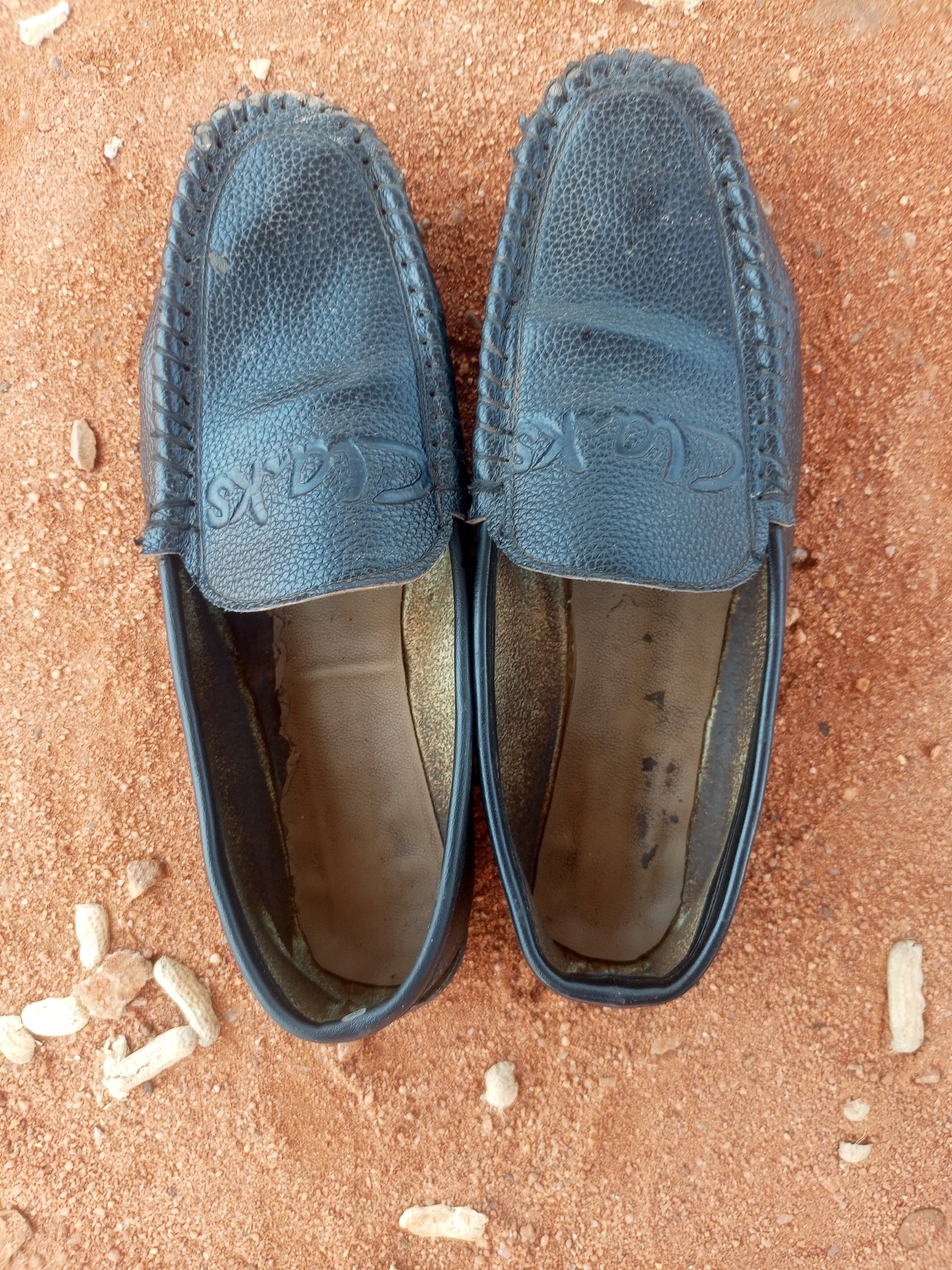 A pair of black leather loafers with a textured finish and decorative stitching are positioned side-by-side on a sandy, reddish-brown surface. The loafers feature embossed branding, which appears to read "CLARKS", on the upper portion of each shoe. The insoles of the loafers show signs of wear, with some scuff marks and discoloration. The surrounding environment is consistent with a dry, outdoor area, possibly in Tempure, Nigeria, as indicated by the reddish sand and scattered small pebbles and what appear to be peanut shells. The lighting suggests it is daytime.