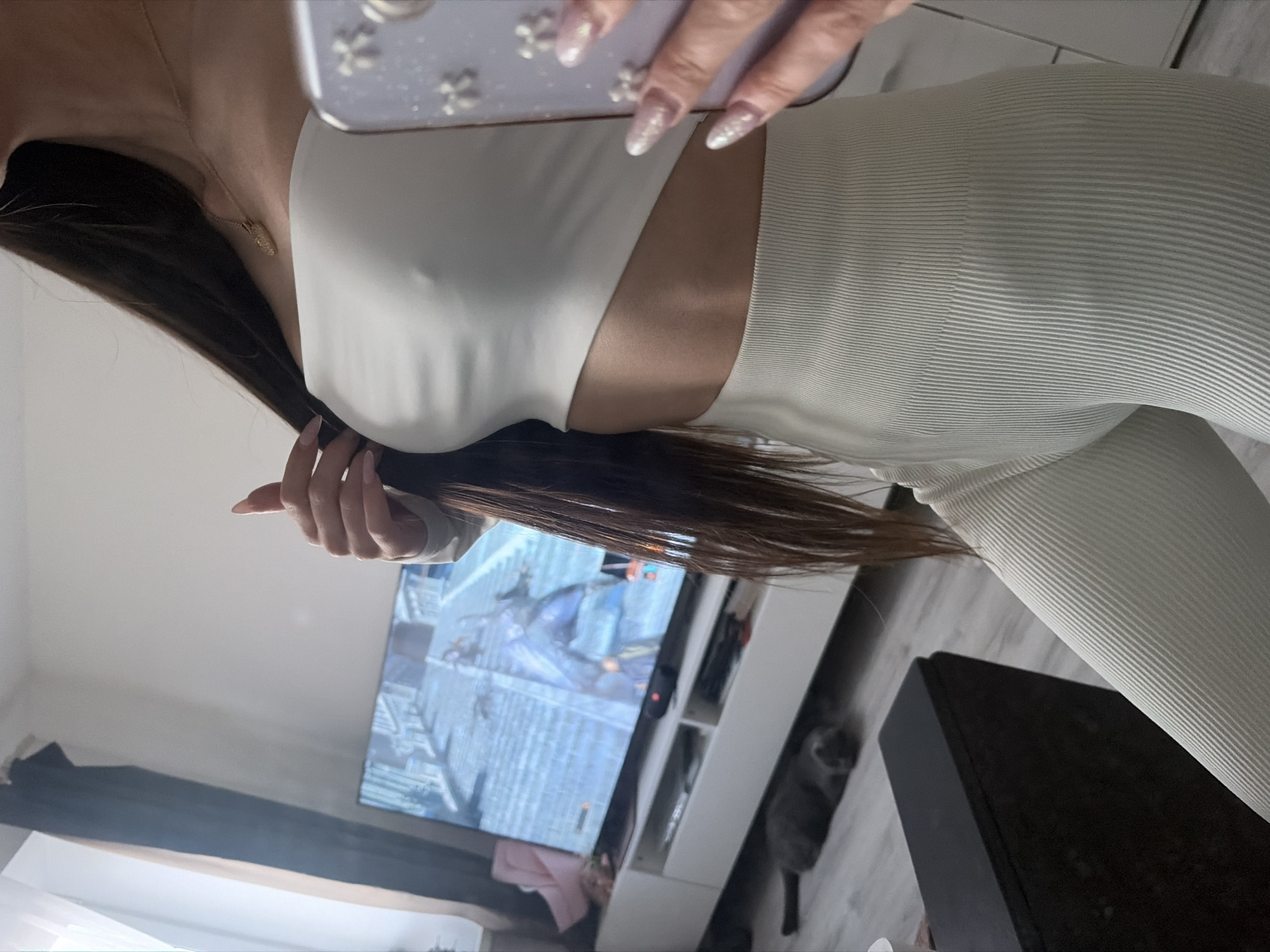 The image depicts a person, likely female, captured from the upper chest to the upper thighs, taking a selfie or mirror picture in an indoor setting. She is wearing a light-colored, ribbed, two-piece outfit, appearing to be a crop top with a cut-out on the side and matching high-waisted leggings or pants. A delicate golden necklace with a small pendant rests on her neck. Her long, dark brown hair cascades around her shoulders, and her left hand is seen touching her hair, revealing long, glittery, manicured nails. Her right hand holds a smartphone with a white case adorned with flower-like embellishments, partially obscuring her face.
In the background, a large flat-screen television displays a scene from what appears to be a video game or fantasy movie, featuring dark figures in a medieval or fantasy setting. Below the TV, a white shelving unit holds various items, including a pink piece of fabric. A grey cat is visible on the light-colored wooden floor to the right of the shelving unit. A dark, rectangular object, possibly a piece of furniture or electronics, is situated on the floor in the foreground. The walls are light-colored, contributing to a bright, modern interior aesthetic of what seems to be a domestic space, likely a living room or bedroom in Saint Petersburg, Russia. No visible text is present in the image.