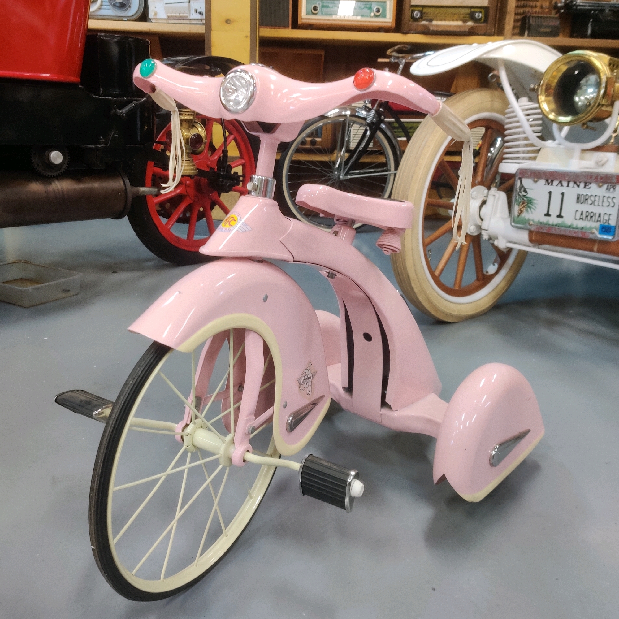 A pink vintage tricycle is the central focus of this image, situated in what appears to be a museum or exhibition space. The tricycle features a bright pink frame, a white seat, and white-rimmed wheels with cream-colored tires. A small red reflector is visible on the handlebars. The tricycle has a headlight and decorative trim. In the background, other vintage items are visible, including what looks like an early automobile or motorcycle with a license plate that reads "MAINE APR 11 HORSELESS CARRIAGE." The floor is a neutral gray. The lighting suggests an indoor environment, possibly with artificial lighting. There are no people present in the image. The context of Yekaterinburg, Russia is not visually supported by the image content.