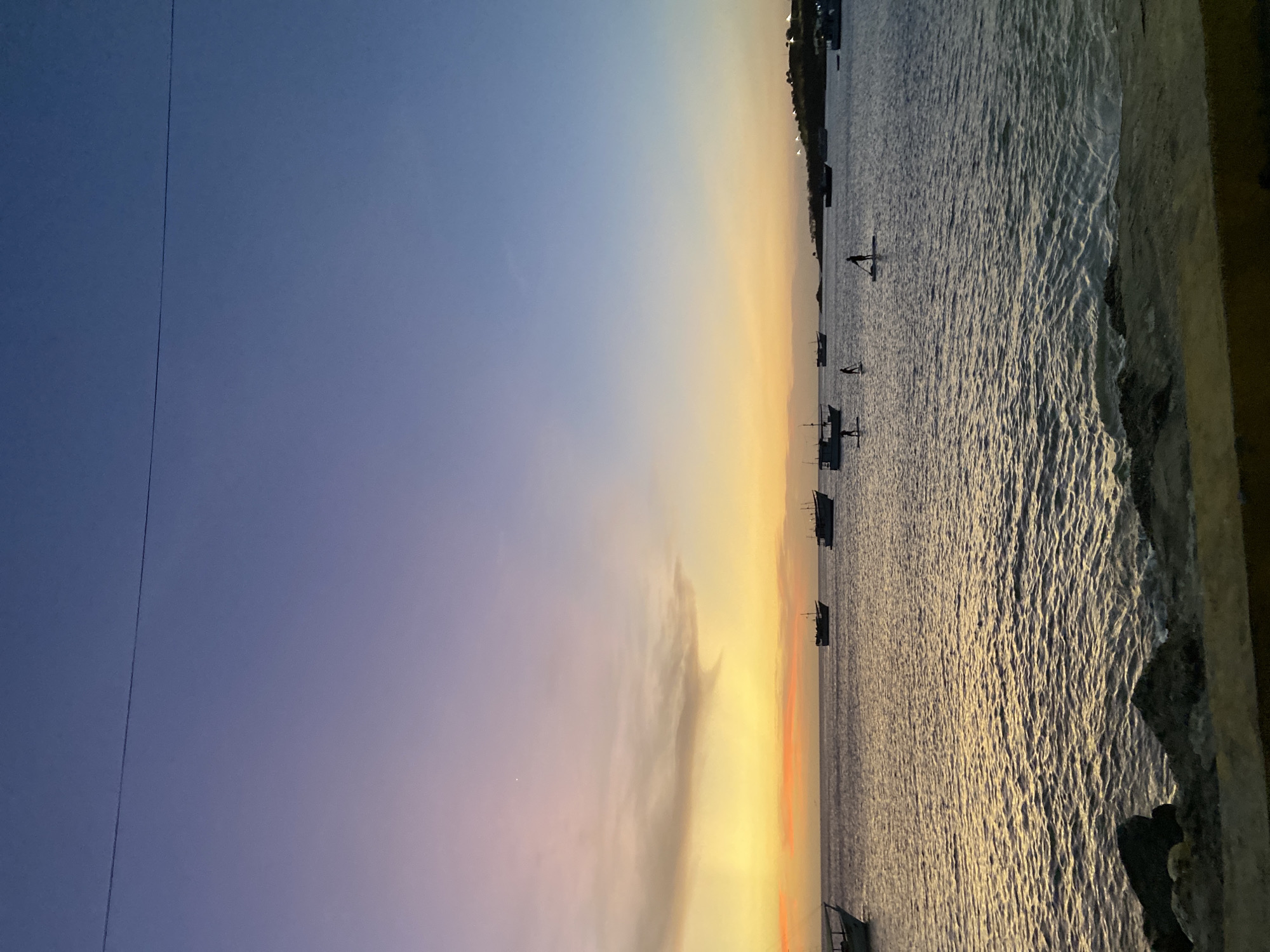 The image depicts a sunset over the ocean in Juan Griego, Venezuela. The sky transitions from blue to purple and yellow near the horizon. Several fishing boats are anchored in the water, and some individuals appear to be standing on paddleboards. A coastline is visible in the distance. A powerline is visible along the upper left side of the image. The foreground shows a part of a concrete structure.