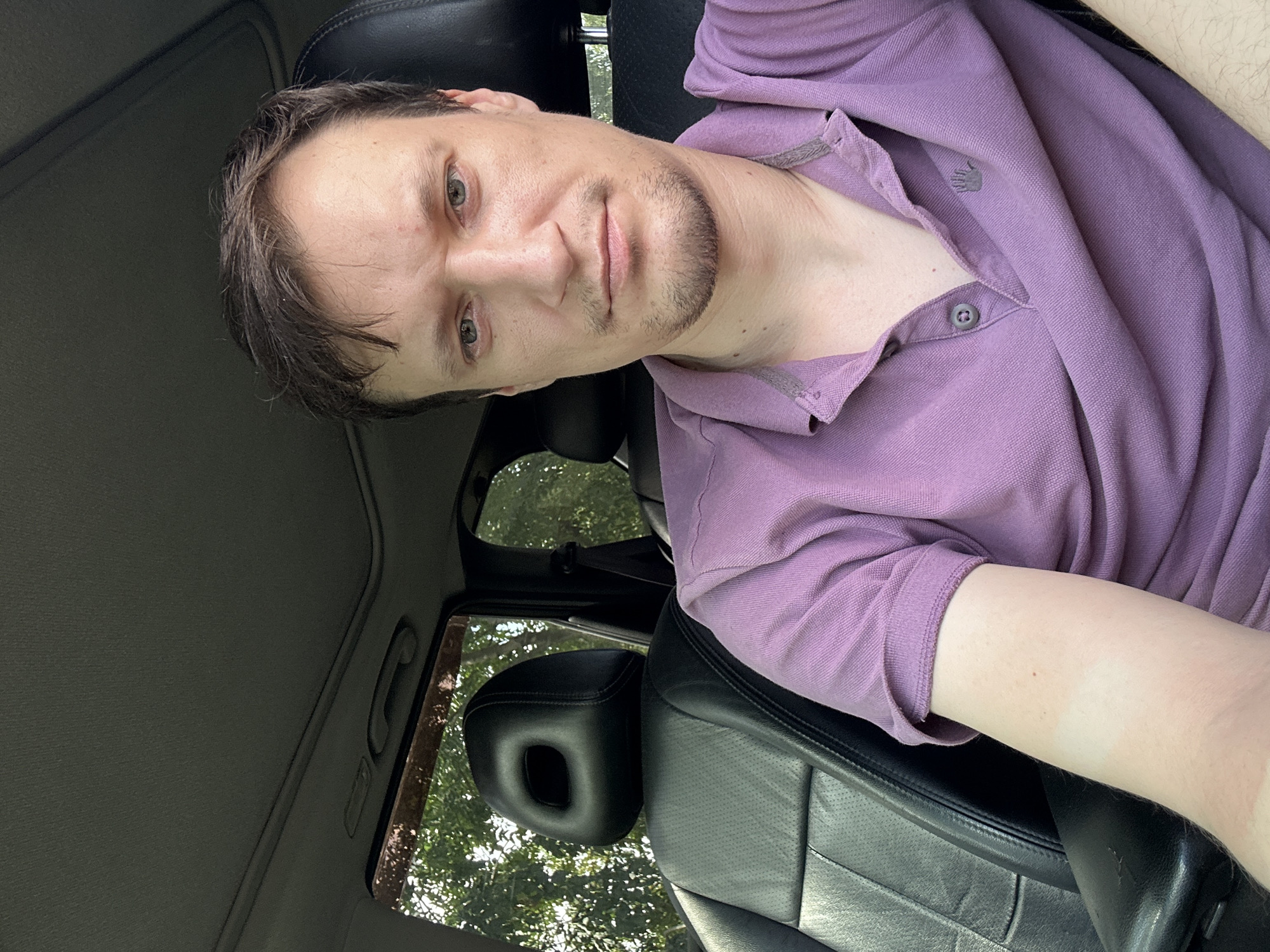 A man is sitting in the driver's seat of a car. He has short brown hair and is wearing a purple polo shirt. The car's interior is visible, including black leather seats and a sunroof. Trees and foliage are visible through the car windows, suggesting an outdoor location, possibly in the city of Makiv, Ukraine, given the provided context. The lighting suggests it is daytime.