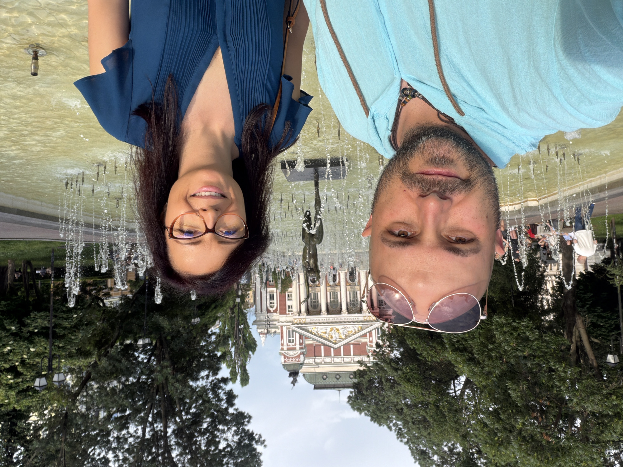 In Sofia, Bulgaria, two individuals, a bald man with sunglasses and a woman wearing glasses, pose for a selfie in front of the Ivan Vazov National Theatre. The man, on the left, wears a light blue hooded sweatshirt and a necklace. The woman, on the right, is dressed in a blue dress. Behind them, a fountain with multiple water jets is active, and in the background, the ornate facade of the National Theatre is visible, surrounded by trees and lampposts. Several other people are seen in the background near the theatre and by the fountain. The lighting suggests it is daytime.