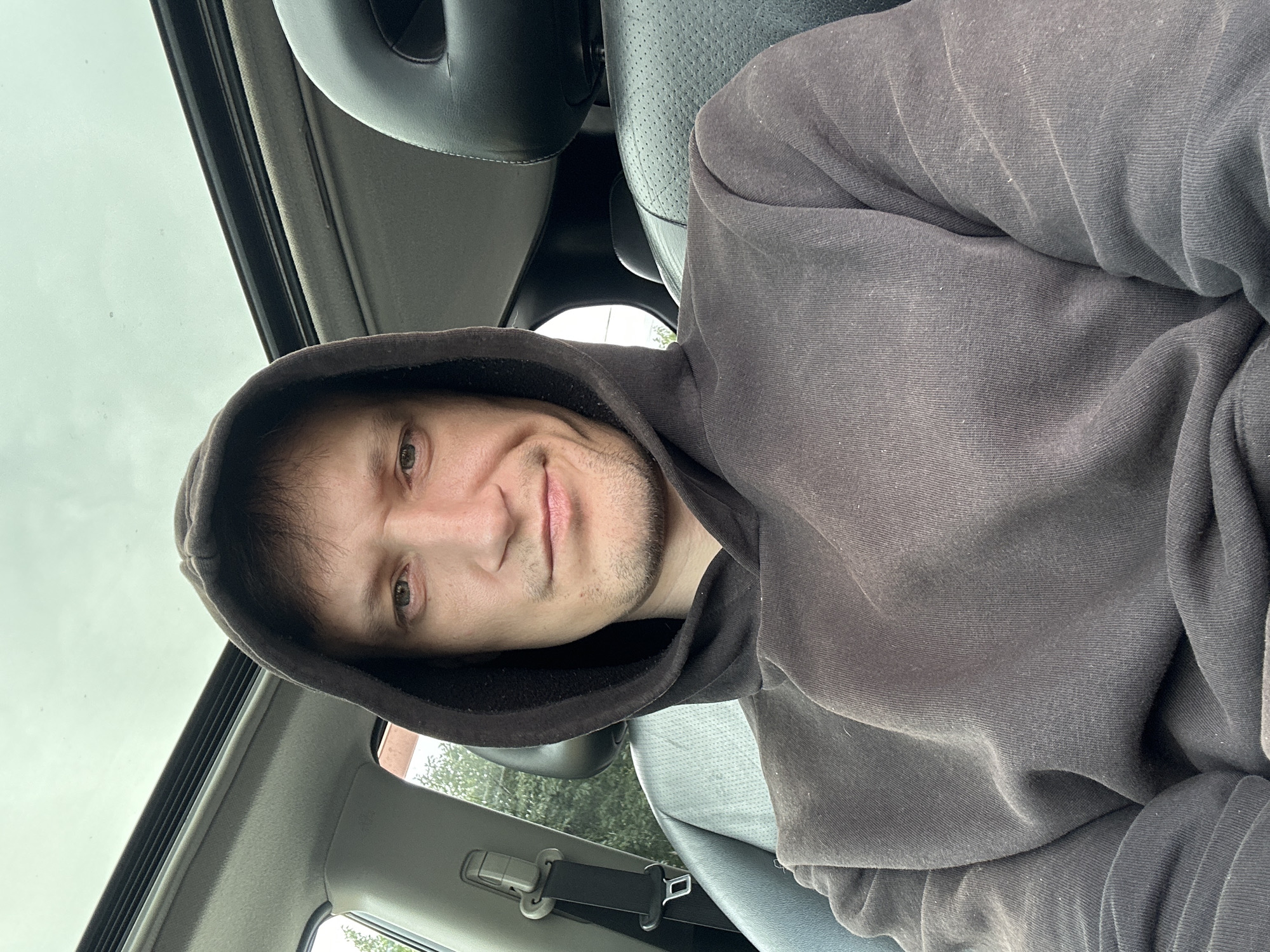 A man wearing a dark hooded sweatshirt is seated in the driver's seat of a car, looking at the camera. The car's sunroof is open, revealing a cloudy sky. The interior of the car includes black leather seats and a seatbelt. The man's face shows a neutral expression. The location is not specified in the image, and it cannot be determined to be in Makiv, Ukraine.