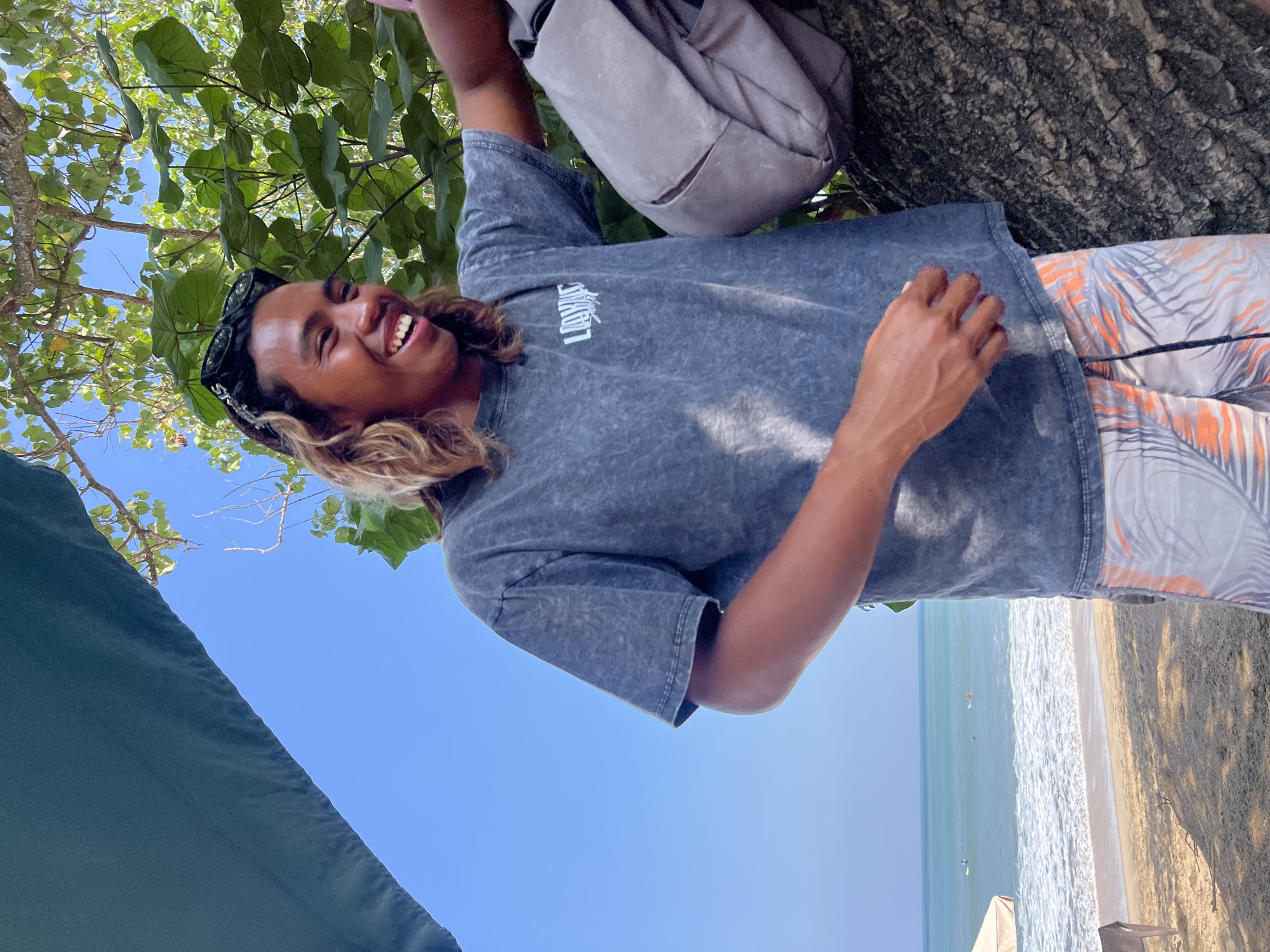 A young man with long, wavy brown hair and sunglasses perched on his head is laughing and looking to his right. He is wearing a dark gray t-shirt with a white logo and patterned shorts featuring orange and gray palm leaves. The man is standing outdoors, leaning against a large tree trunk with rough bark. Behind him, a calm, turquoise ocean meets a sandy beach, with several surfers visible in the water. The sky is a clear, bright blue. A portion of a teal-colored canopy is visible in the upper left corner of the image. The scene appears to be Kuta, Indonesia, given the tropical beach setting and surf activity.