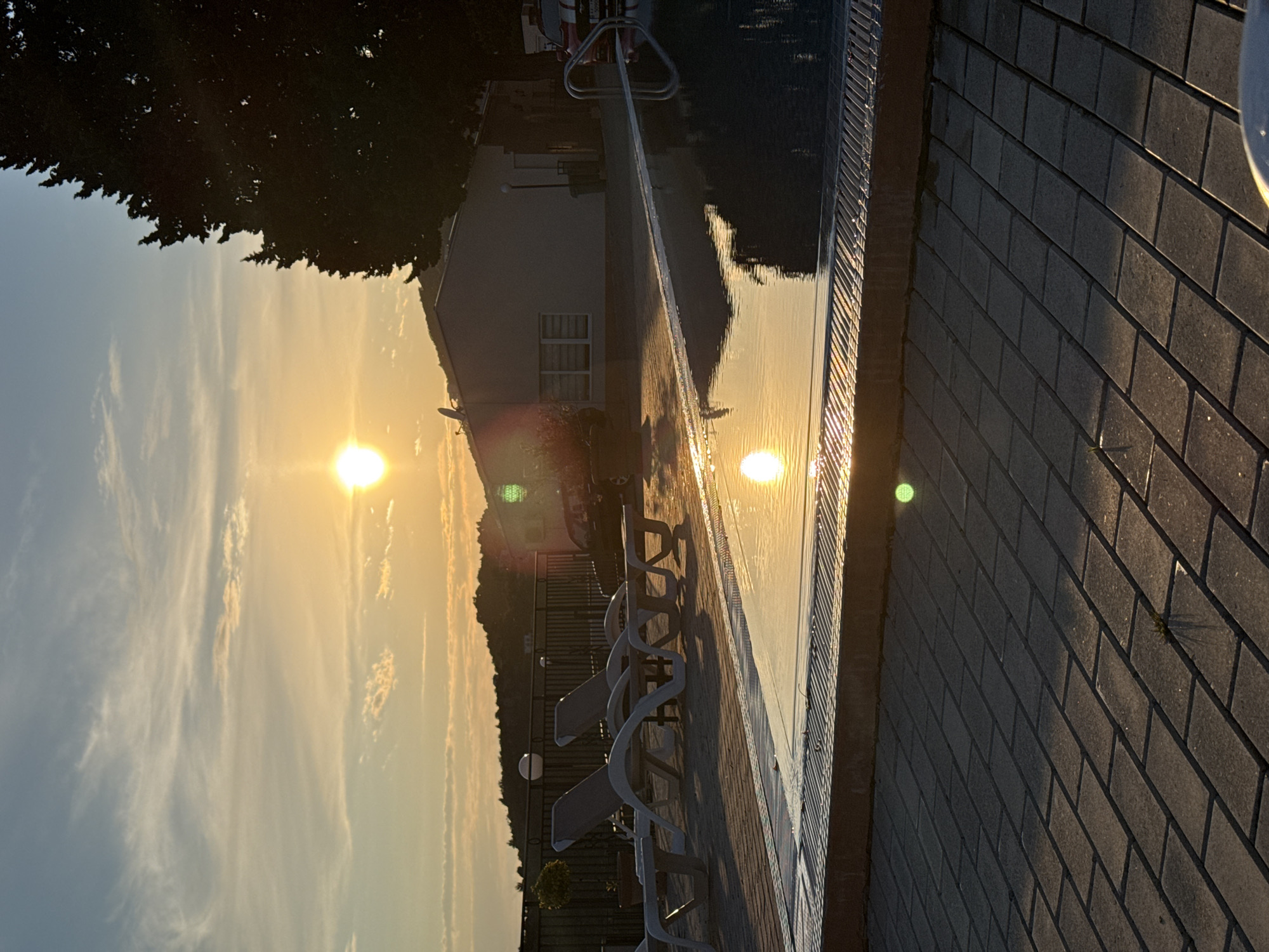 The image depicts a serene evening scene at a pool in Šušanj, Montenegro. The sun is setting, casting a bright glow and warm colors across the sky and reflecting in the pool water. Adjacent to the pool, lounge chairs sit on a paved surface. A building, possibly a hotel or residence, stands in the background. Trees and hills complete the landscape.