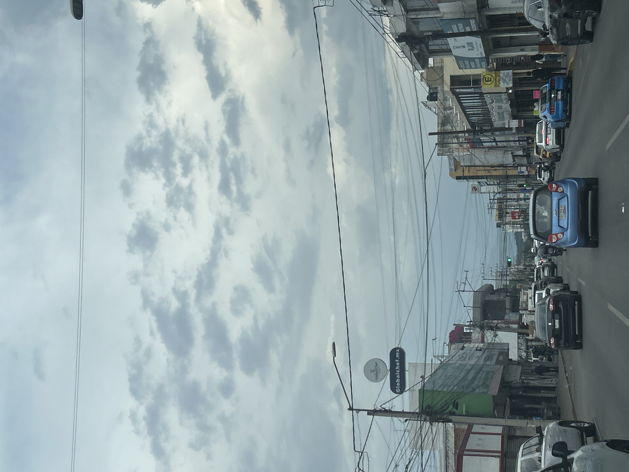 The image captures a street scene in Puebla, Mexico. The sky is overcast with a mix of grey and white clouds. Buildings line the right side of the street, with a variety of signs visible. There are multiple cars on the street, including compact cars, SUVs, and sedans. Power lines and utility poles run along the right side of the street. A sign that reads "globalchef.mx" is visible on one of the poles. The traffic signal is green.