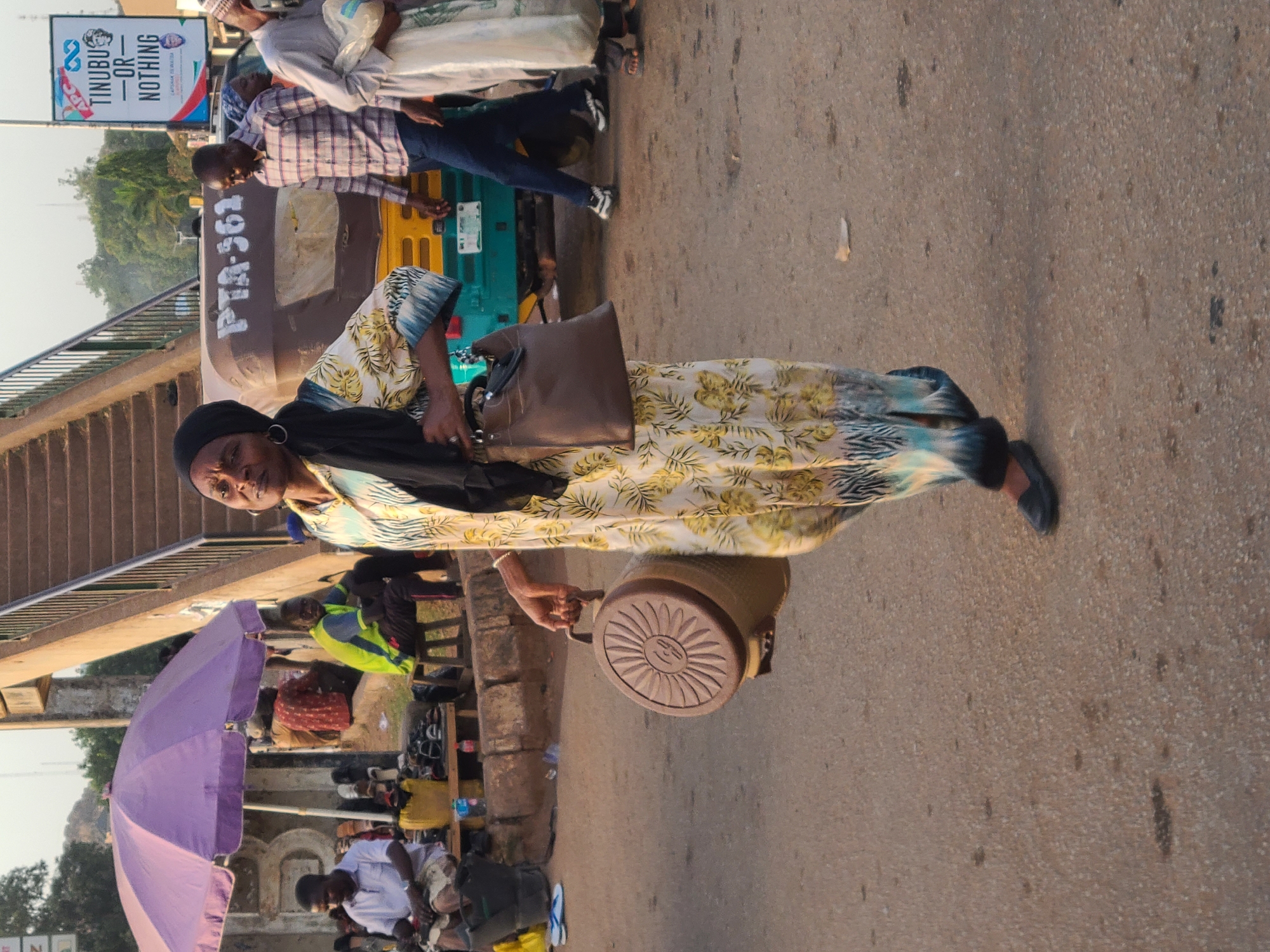 The image shows a woman walking on a street in Jos, Nigeria, near a pedestrian overpass. She is carrying a brown handbag and a cooler with a decorative sun design on the lid. She is wearing a floral-patterned dress, black shoes, and a black head covering. Behind her, there is a brown and green vehicle, possibly an auto rickshaw or taxi. A political banner that reads "TINUBU -OR- NOTHING" is visible above the vehicle. Several people are visible in the background, some seated under a purple umbrella, likely vendors. The street appears to be busy.