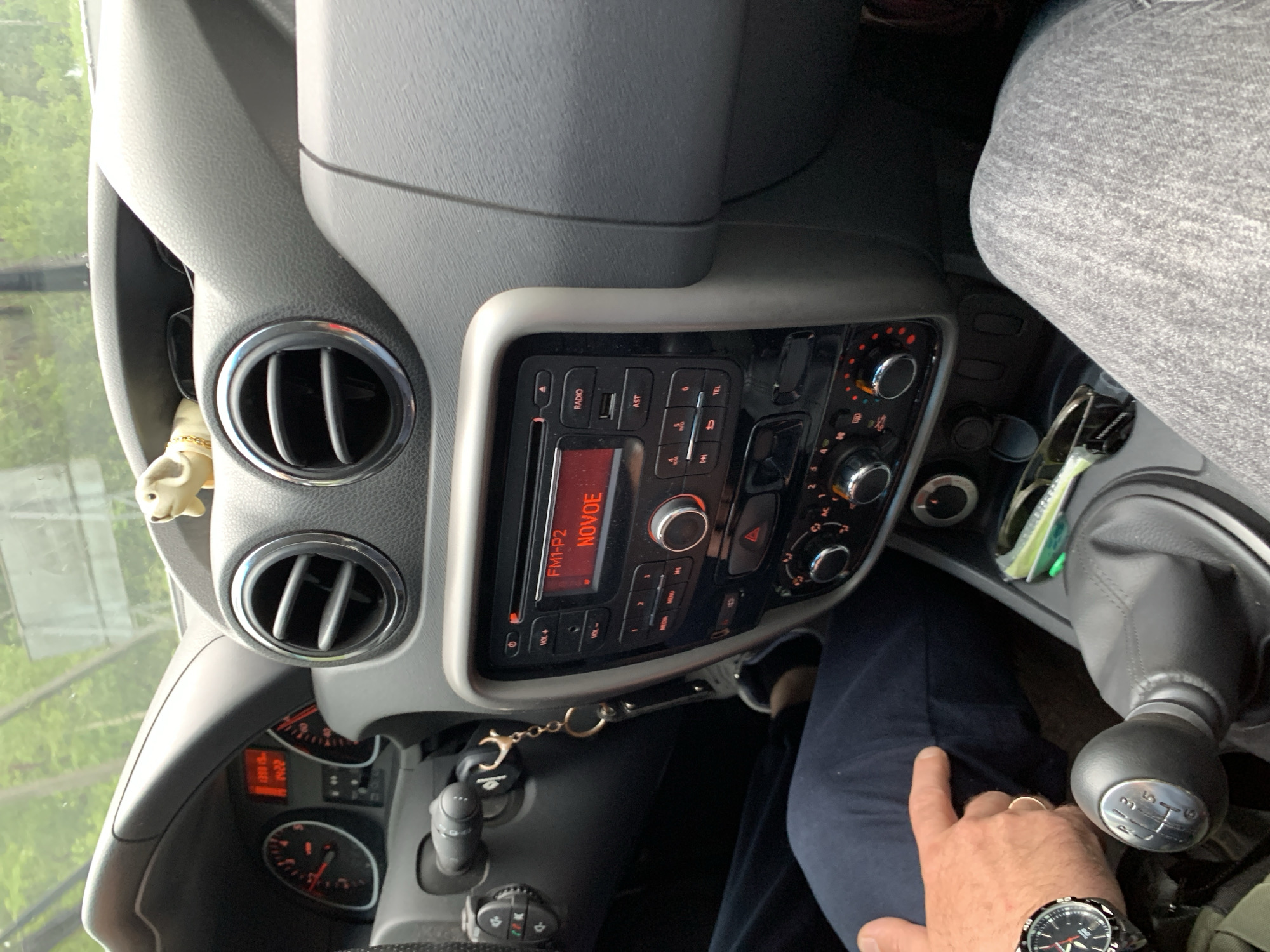 The image shows the interior of a car, likely a Dacia Duster, viewed from the driver's perspective. A person's hand, wearing a watch and a ring, is visible in the lower right corner, resting on their dark blue pants. The dashboard features the car's radio display, showing "FM1-P2 NOVOE," along with various buttons and knobs for controlling the audio system and climate. Two air vents are also visible on the dashboard. The gear shift is prominently displayed, indicating a manual transmission. The scene appears to be set during the daytime, with natural light filtering into the car, suggesting an outdoor setting, possibly in a rural area given the location context of Мядзельскі сельскі Савет, Belarus. The overall impression is one of a mundane car interior, with the focus on the functional elements of the vehicle and a hint of the driver's presence.