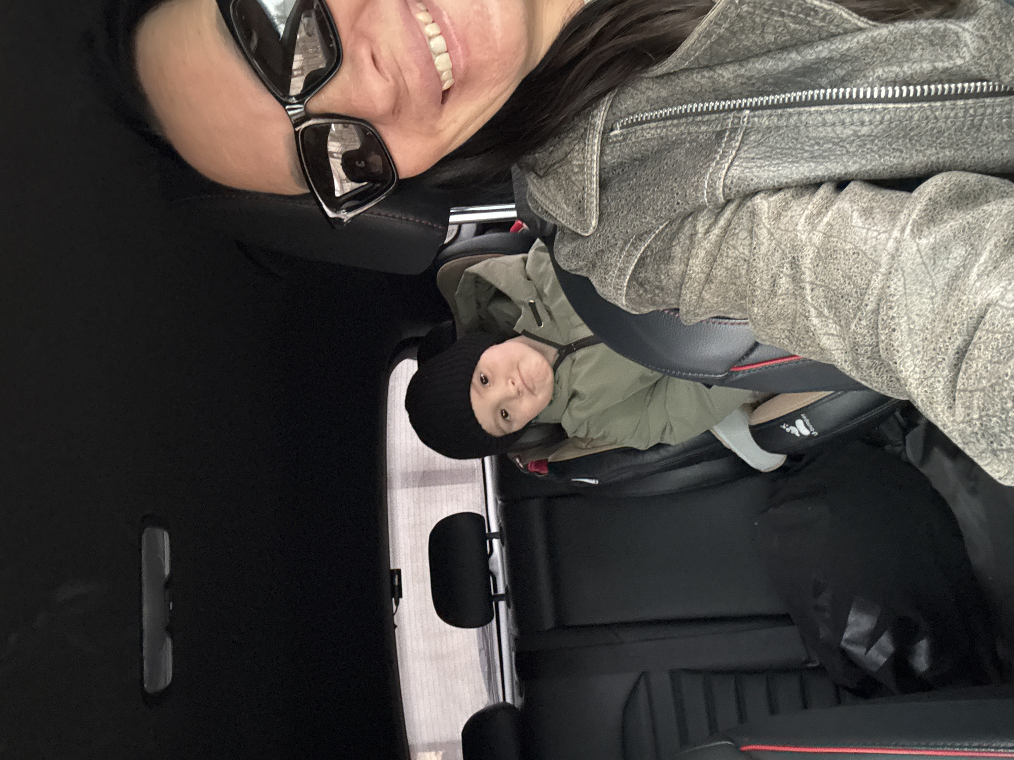 A selfie inside a car shows a woman with sunglasses and a grey leather jacket in the front passenger seat. A young child wearing a black hat and an olive-green jacket is in a car seat in the back. The interior of the car is black with red trim. A reflection of what appears to be the side of a building and parking spaces is visible in the car window in the background, suggestive of an urban or suburban environment. Based on the query request for the location of Chaikovsky, Russia, it can be assumed that this photograph was taken there.