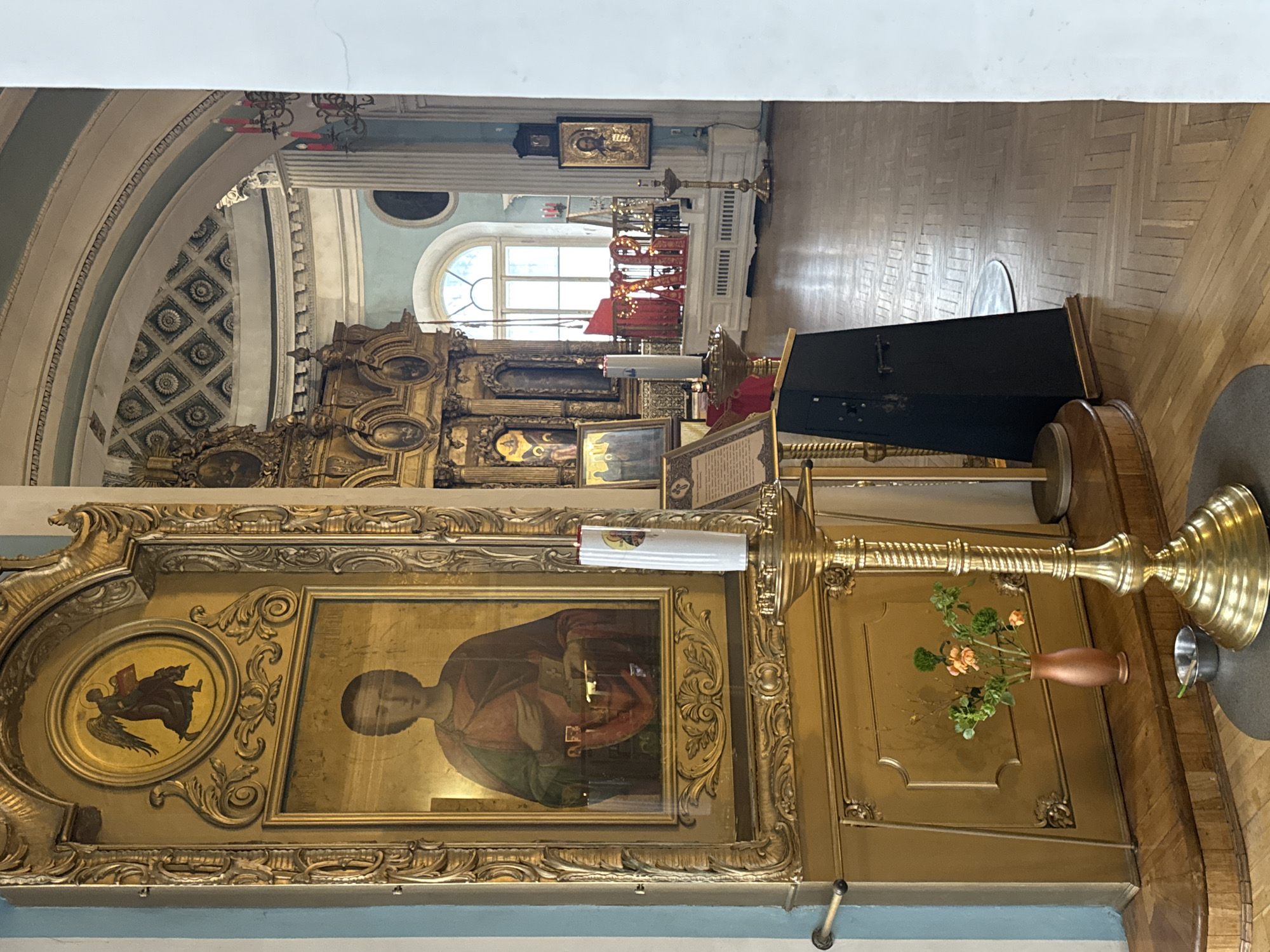 This is a vertical interior shot of an Orthodox Christian church or cathedral in Saint Petersburg, Russia. The scene is dominated by religious artifacts and architectural details.
In the immediate foreground and to the right, a large, ornate golden candelabra or candle stand with intricate spiral carvings is visible. Beside it, a small brown vase holds a bouquet of light orange blossoms and green foliage. A small silver-colored metal cup or container rests near the base of the candelabra.
The central focus is a richly decorated iconostasis, an altar screen adorned with numerous religious icons set in elaborate golden frames. A prominent icon on the screen depicts a figure, possibly a saint, holding a book. Above and to the left of this main figure, a smaller circular icon shows a winged figure, likely an angel. The iconostasis features extensive gold detailing and carved scrollwork. A white sheet or banner with illegible text is attached to the candelabra, partially obscuring the iconostasis.
In the mid-ground, behind the iconostasis, an arched window allows natural light into the space, revealing glimpses of the exterior. Further back, more elements of the church's interior architecture can be seen, including columns, another icon (perhaps of Christ) on a wall, and a piece of red fabric or banner.
The ceiling features decorative arched structures and coffers with circular rosette patterns. A circular window is visible higher up on a pale blue-grey wall. The floor is made of light-colored wooden parquet, laid in a herringbone pattern, stretching into the background.
No specific activity is taking place; the scene captures the static and reverent ambiance of the church interior during what appears to be daytime. The overall impression is one of solemnity and historical grandeur.