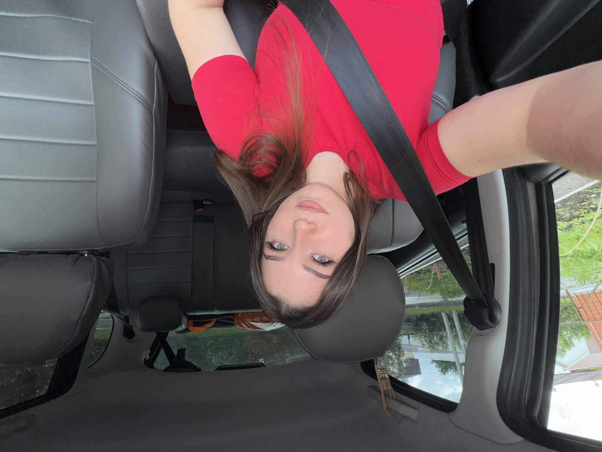 A young woman with brown hair and green eyes, wearing a red t-shirt and a black seatbelt, sits in the passenger seat of a car. She is looking at the camera. The interior of the car, including the grey seats and a portion of the dashboard, is visible. Outside the car window, residential buildings and greenery can be seen, consistent with a suburban or semi-rural area. The image appears to have been taken during the daytime. The location is identified as Perm, Russia.