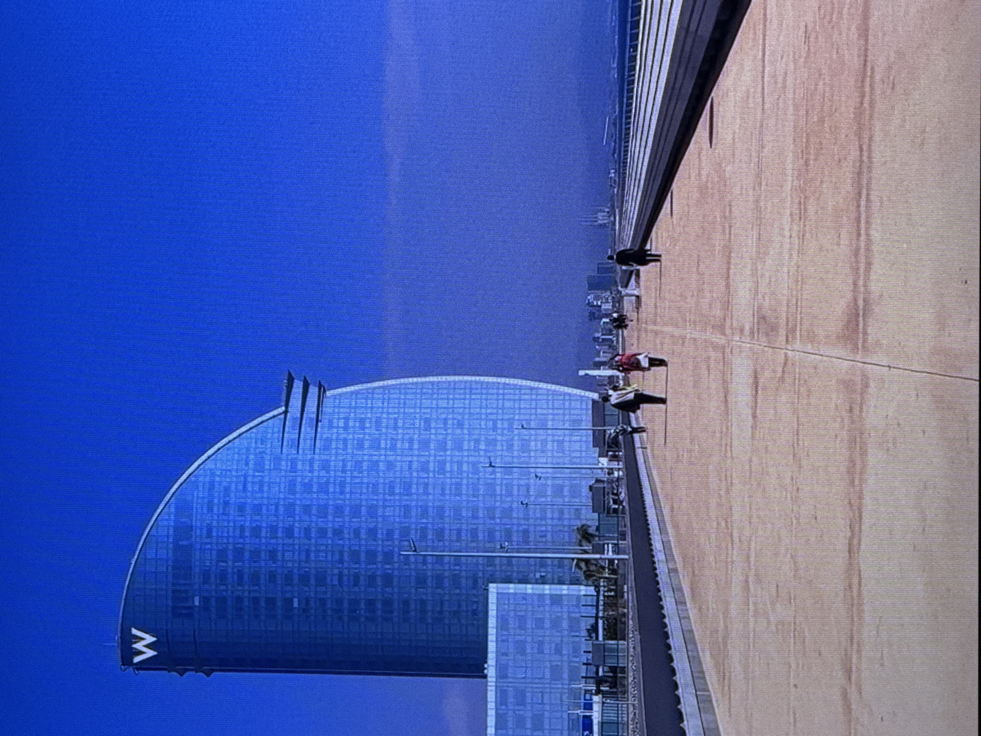 The image shows a wide pedestrian walkway with a sandy-colored surface, leading towards a city skyline in the distance under a clear blue sky. On the left side of the walkway, a tall, curved, glass-covered building with a prominent "W" logo at its peak dominates the scene. Further back, another smaller glass building is visible. Several lampposts are spaced along the walkway. Four people are seen walking on the walkway, appearing to be heading away from the viewer. The individual closest to the viewer on the right is wearing a dark coat and carrying a bag. To their left, another person in a red jacket and white scarf is walking. Two more figures are further ahead on the walkway, also walking away. The overall environment appears to be urban, possibly a waterfront promenade. The city skyline in the distance includes several structures, and a body of water is visible beyond the city. There is no indication that this scene is in Dubai, United Arab Emirates.