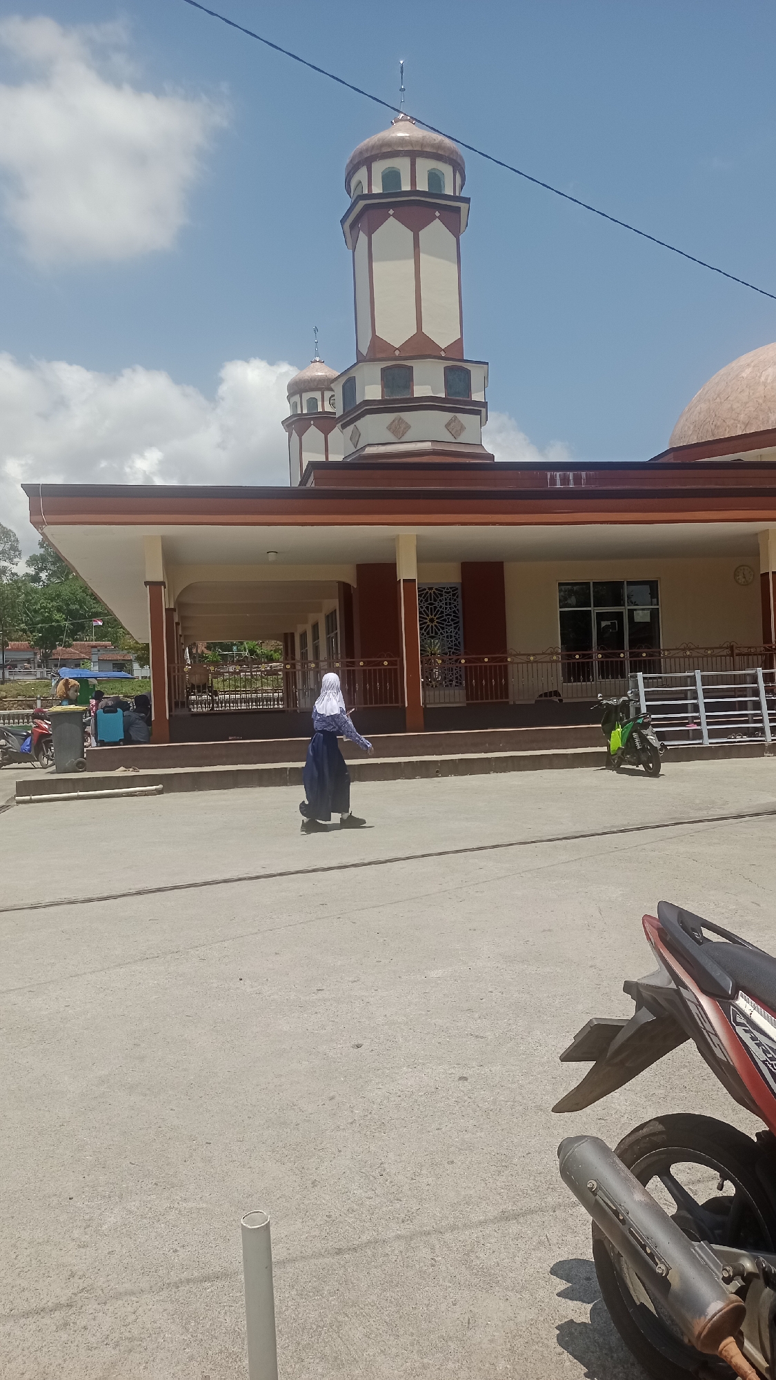 The image captures a daytime scene outside a mosque in Pakemitan, Indonesia. A person dressed in a blue hijab and dark blue attire walks across a paved area in the foreground. Behind them, the mosque features a prominent dome and two tall minarets, with the main building having a covered walkway with columns and windows. Several motorcycles are parked near the entrance of the mosque, and in the background, there are trees and a glimpse of other buildings. The sky is blue with scattered clouds.