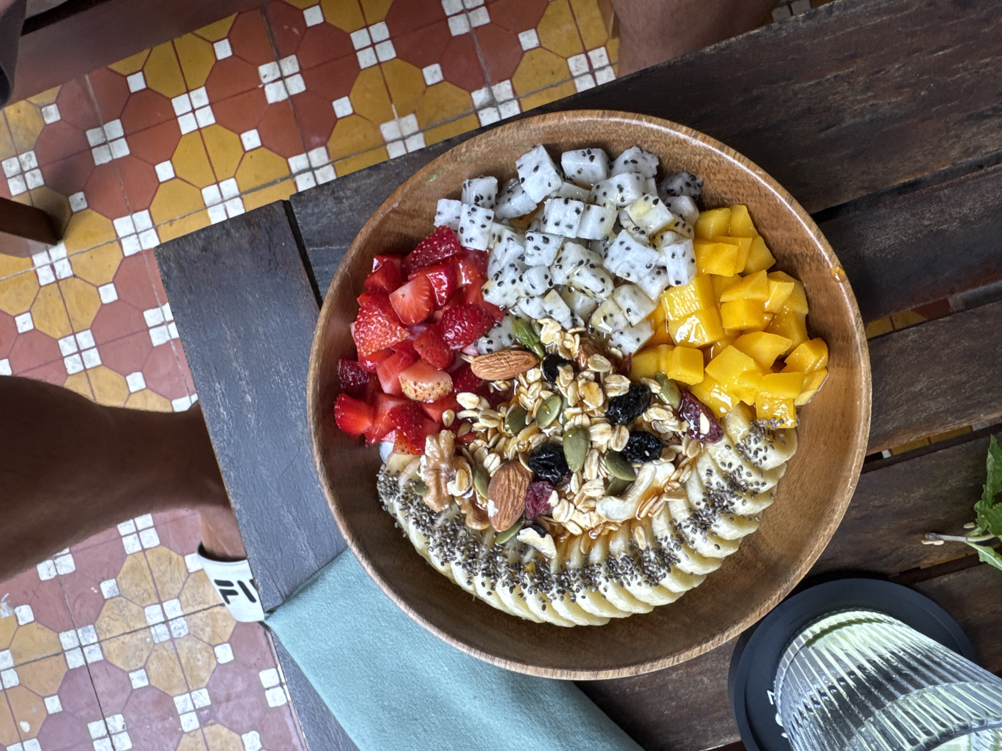 The image captures a wooden bowl filled with a vibrant array of fruits, grains, and seeds, arranged artfully. The bowl contains diced dragon fruit, sliced strawberries, cubed mango, granola with mixed nuts and seeds, and sliced bananas topped with chia seeds. It is likely a smoothie bowl. The bowl rests on a dark wooden table next to a blue napkin and a glass of water. The table sits on a patterned tile floor. A leg wearing a white sandal is visible to the left of the table.
While the image does not explicitly state that it was taken in Đà Nẵng, Vietnam, the presence of dragon fruit (a commonly grown and consumed fruit in Vietnam) increases the probability it could be there. The tile floor pattern is also consistent with styles seen in Southeast Asia. Without additional contextual information, it is impossible to definitively confirm the location in Đà Nẵng.