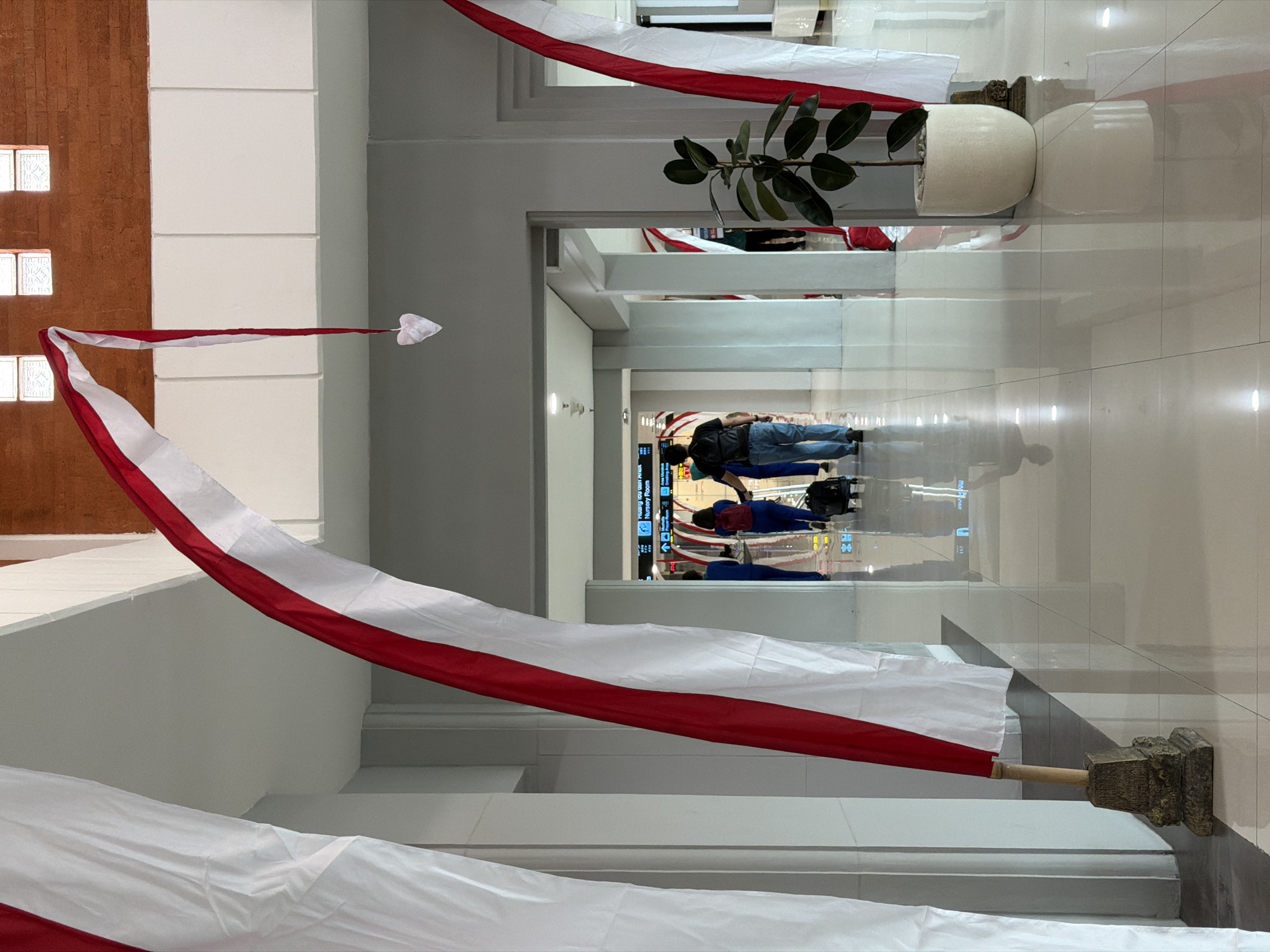 An indoor hallway is decorated with large, flowing Indonesian flags and a small heart ornament. The flags are red and white, with wooden poles resting on ornate bases. The hallway has a tiled floor that reflects the ambient light from ceiling fixtures. In the background, several people are walking, some pulling luggage. A potted plant with large green leaves sits to the right of the frame. The architecture suggests a public building, possibly an airport or a similar transit hub. The lighting is bright and diffused.