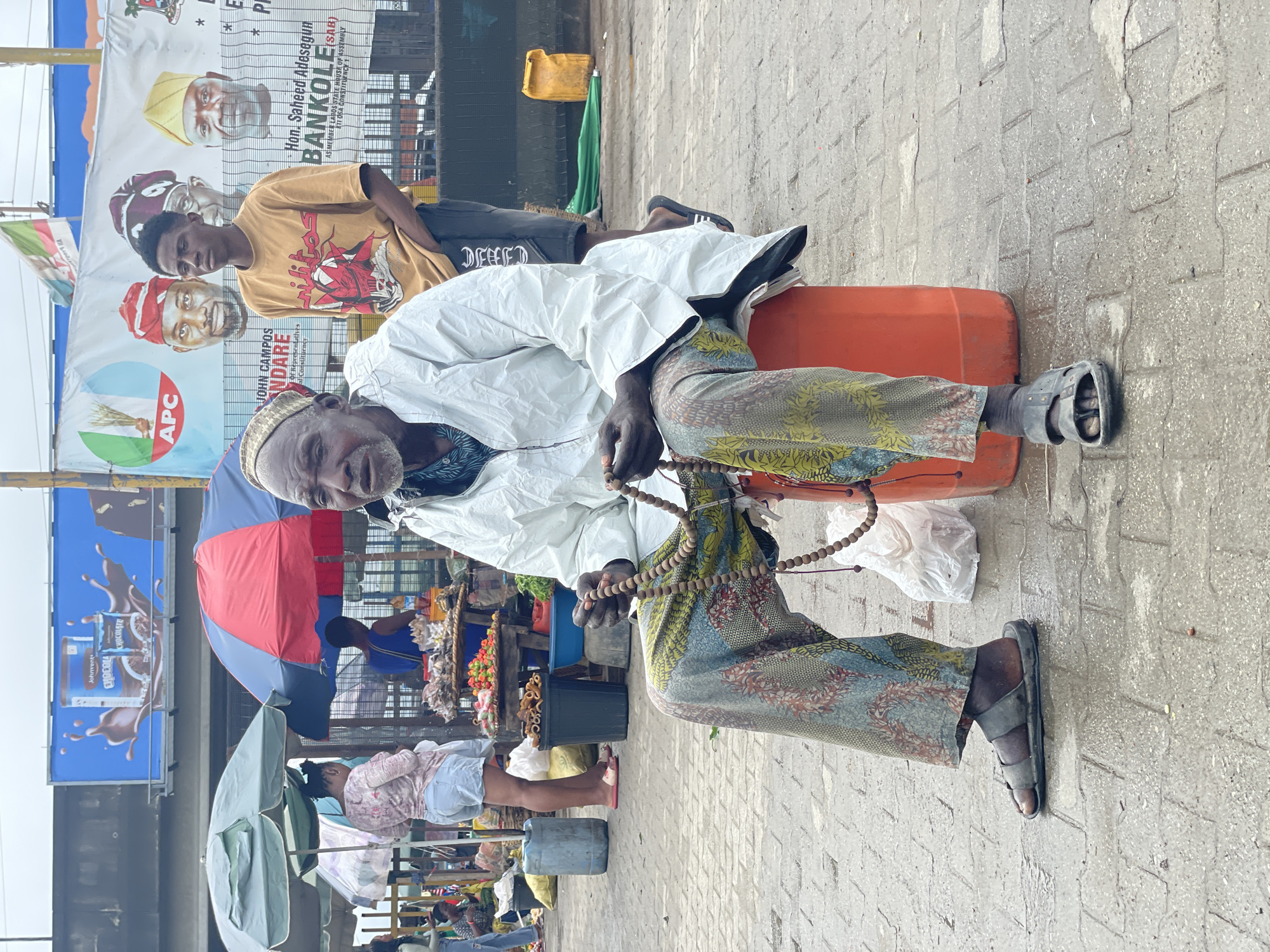 A man in traditional Nigerian attire, including a patterned wrapper and a white robe, sits on an orange plastic barrel at a market stall in Ajah, Nigeria. He is wearing a religious head covering and holding a string of prayer beads in his hands. A young man stands to his right, looking away from the camera. In the background, a large banner displays political figures and text in English and possibly Yoruba. Various market stalls with umbrellas, produce, and other goods are visible. The ground is wet, suggesting recent rain.