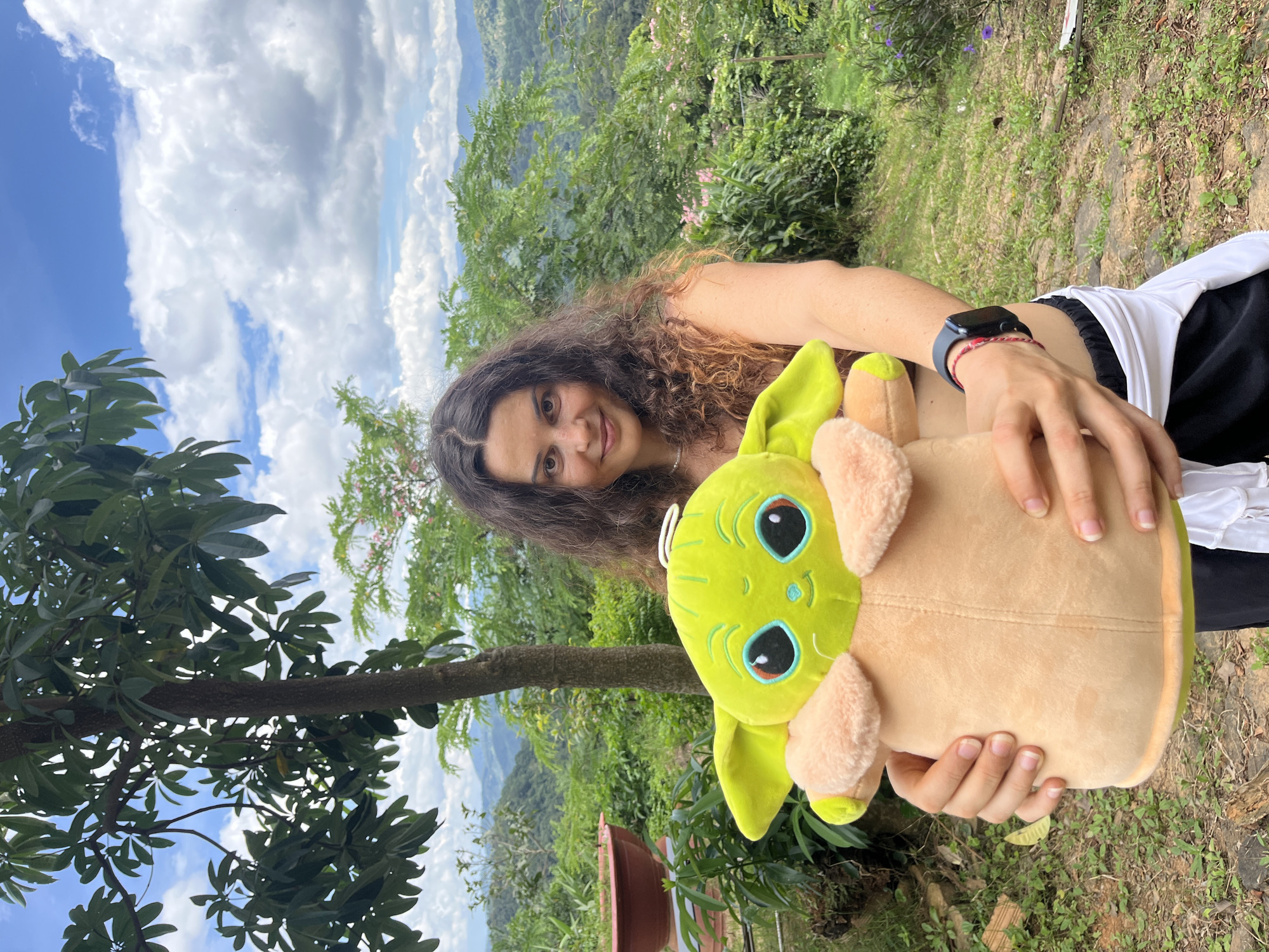 A young woman with long, curly brown hair stands outdoors, holding a large, lime-green plush toy resembling a baby Yoda. She is wearing a dark bikini top and black athletic shorts with a white wrap. Her arms are extended forward, cradling the toy. The background features lush green vegetation, rolling hills covered in trees, and a cloudy blue sky, suggestive of a mountainous or elevated rural landscape, possibly in Sơn Điền, Vietnam, known for its natural beauty. A tree trunk and foliage are visible to her left, and a large brown pot is also present on the left side of the frame.