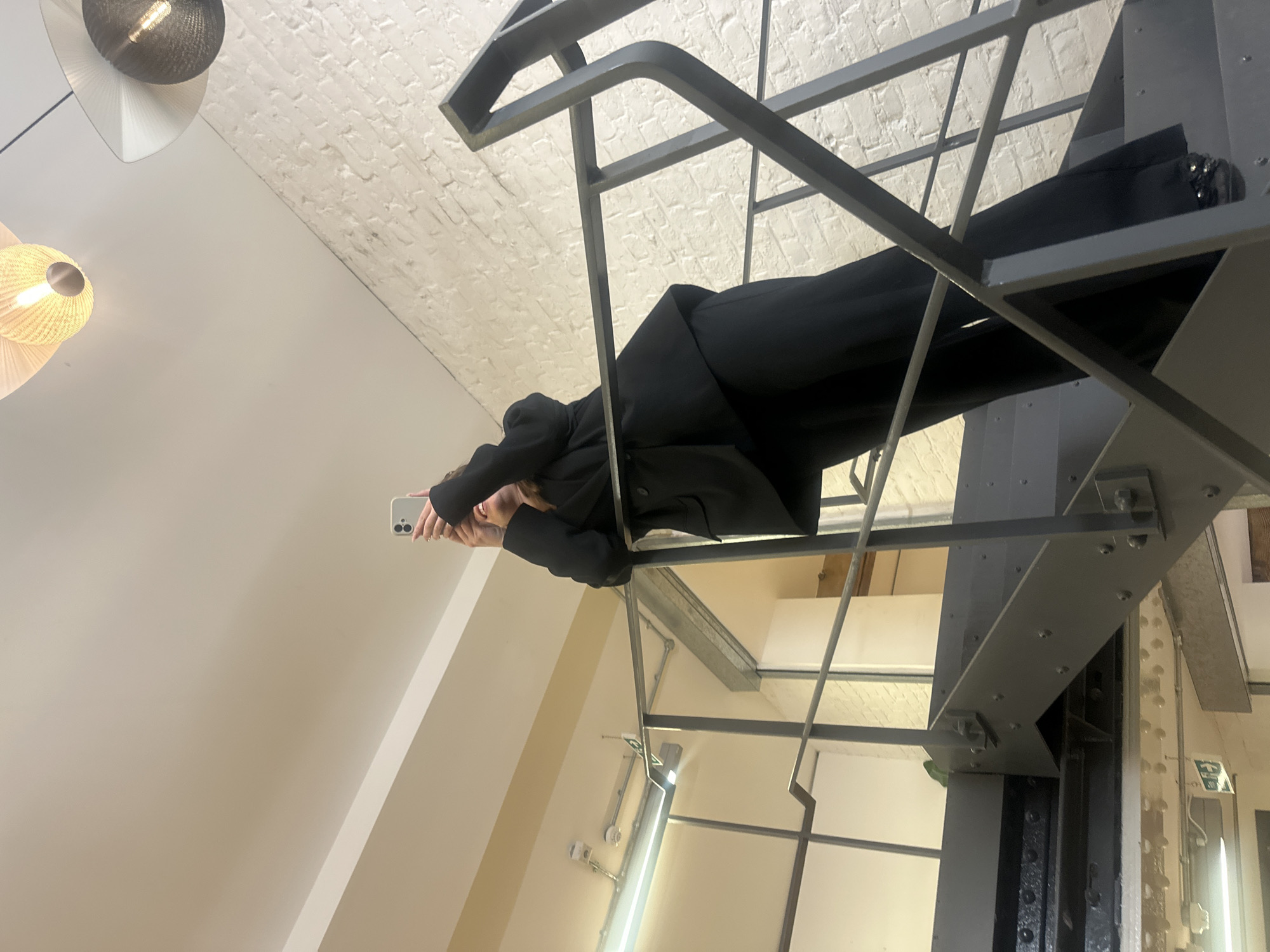 An interior photograph captures a person engaged with their smartphone within a modern, industrial-style architectural space. The image is taken from a slightly low angle, emphasizing the dynamic diagonal lines of a prominent dark grey metal staircase or structural framework that traverses the scene from the lower right to the upper left.
The subject, positioned centrally within the metal framework, is dressed in all-black attire, appearing to wear a blazer or jacket and trousers. Their hands are raised, holding a light-colored smartphone (resembling an iPhone) towards their face, indicating they are likely taking a selfie or recording a video. Their face is largely obscured by the phone.
The background reveals a textured white brick ceiling or upper wall, contrasted with smooth cream-colored walls. To the left, two contemporary pendant light fixtures are visible, featuring distinct spherical shades and conical diffusers. Further into the background, elements of the building's industrial design are apparent, including a large riveted metal column, exposed conduit pipes, and a bright linear light strip. A green emergency exit sign is also partially visible on a wall in the distance.
The setting suggests a renovated urban building, possibly an art gallery, modern office, or public space, characterized by its blend of raw industrial materials and contemporary finishes, fitting the context of London, United Kingdom. The artificial lighting indicates an indoor scene. The image presents a casual moment of interaction with technology within a distinctive architectural environment.