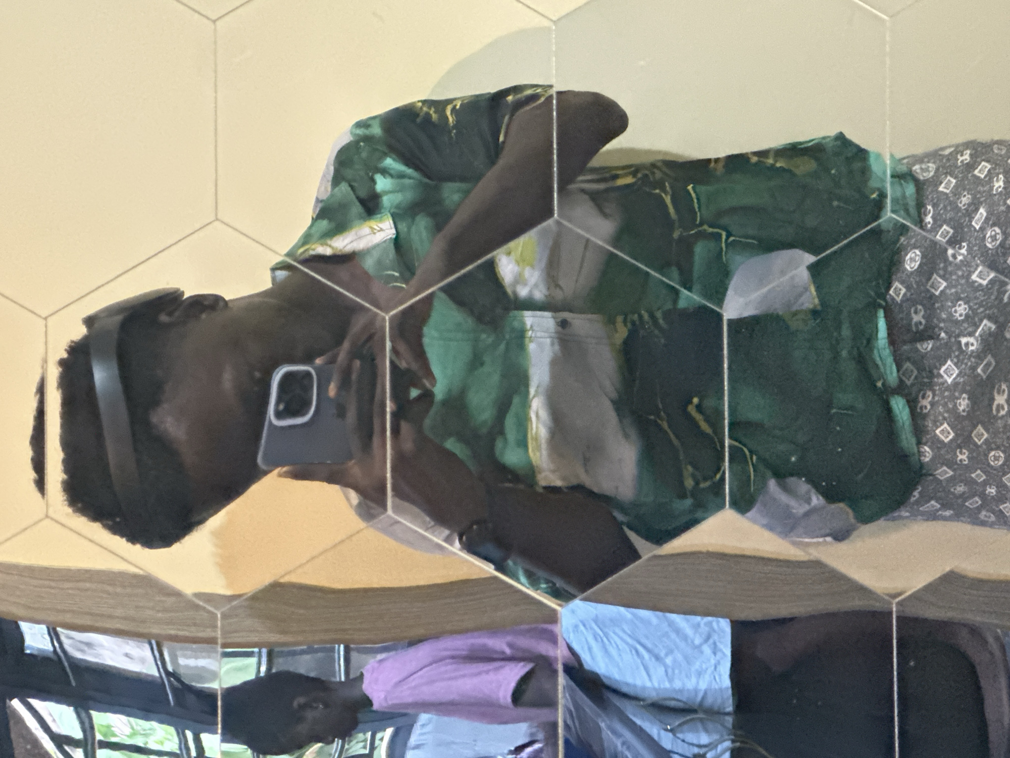 A young Black male, wearing a black headband, a green and white patterned short-sleeved button-up shirt, and gray patterned shorts, is taking a selfie in a hexagonal mirror. The mirror reflects a person in a pink t-shirt and blue shorts to the left, and a window with greenery outside. The background wall is light yellow. The scene appears to be indoors, possibly in a home or a commercial establishment, located in the city of Ajara, Nigeria.