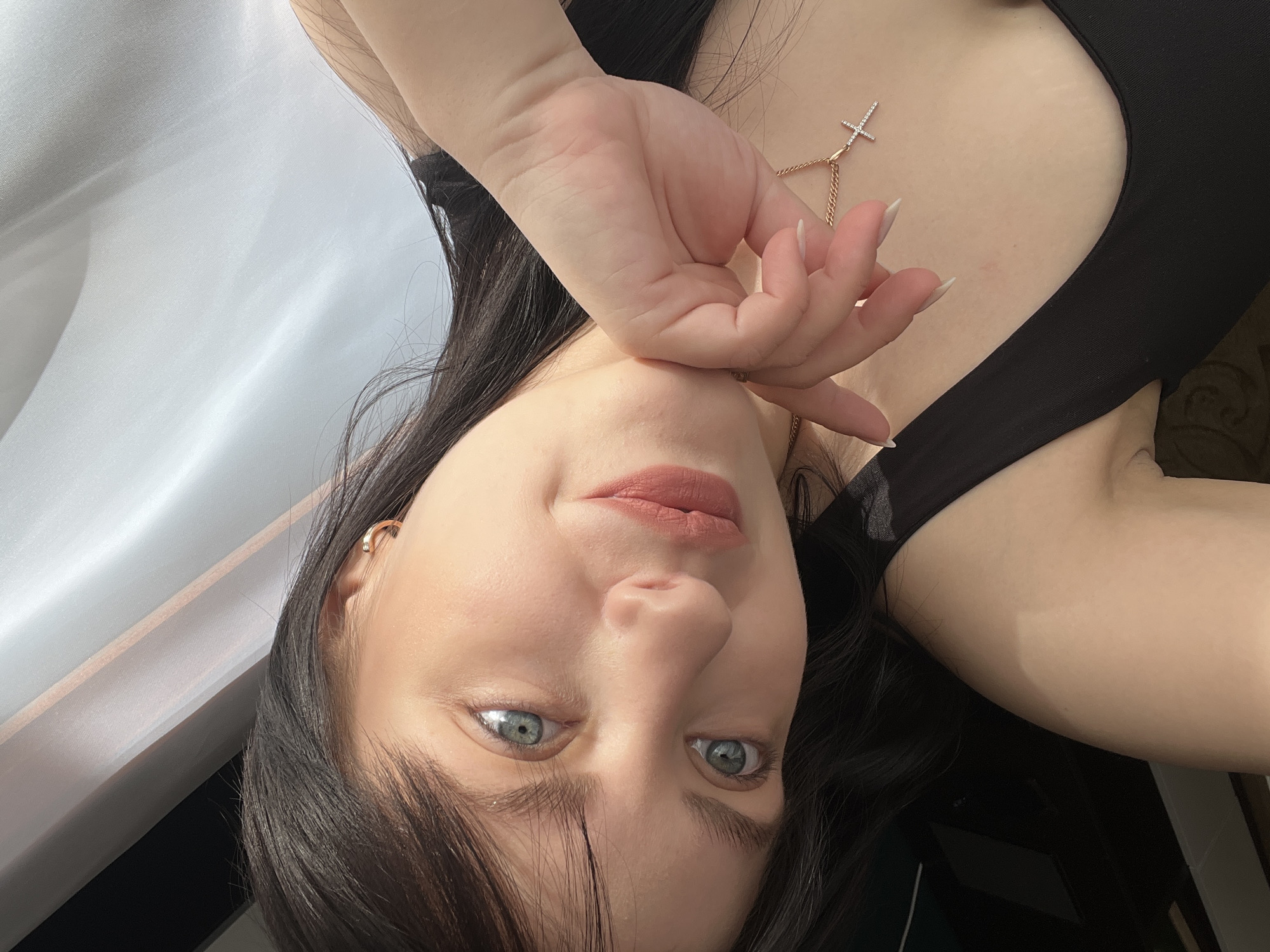 A woman with dark hair, light skin, and blue eyes is posing for a photograph indoors. She is wearing a black top and a delicate gold necklace with a cross pendant. Her left hand is near her face. The background includes a white curtain. The photo appears to have been taken in Марковский, Russia.