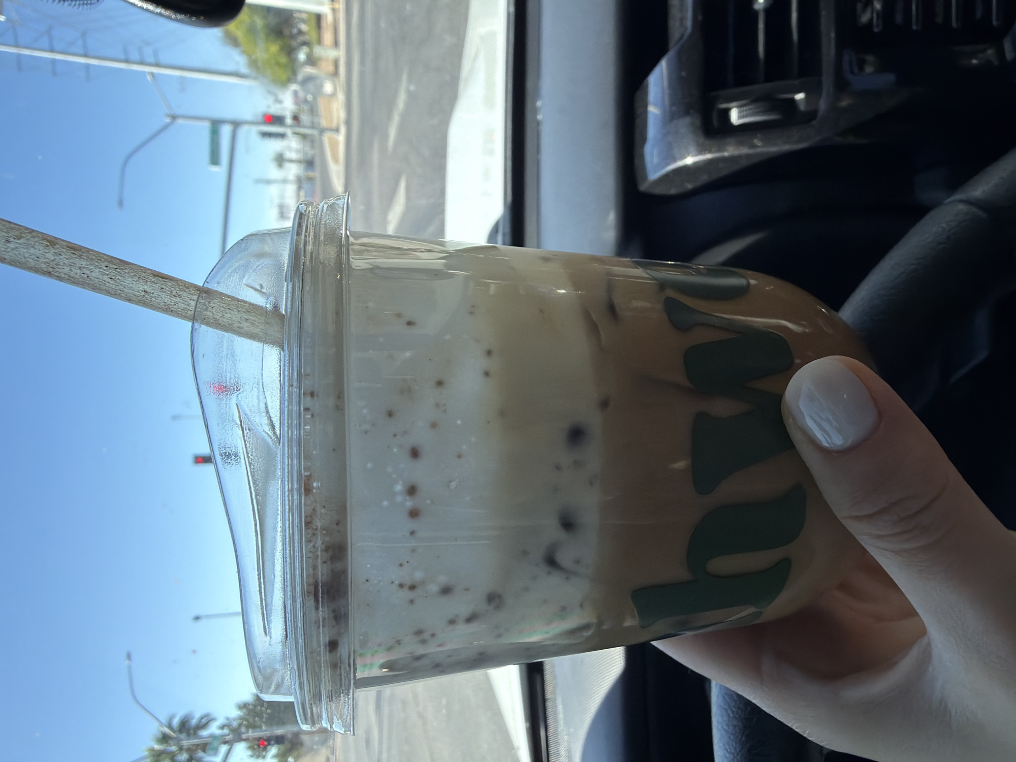 A cup of iced coffee is held in a hand with a white fingernail against the backdrop of a car interior and the city of North Las Vegas, United States of America. The transparent plastic cup has a lid and a straw, and contains a layered drink, likely coffee with milk and ice. The coffee cup's brand is visible. The car's interior includes a black steering wheel and air vents. Outside the car window, the sky is bright blue, and streetlights, palm trees, and traffic signals are visible.