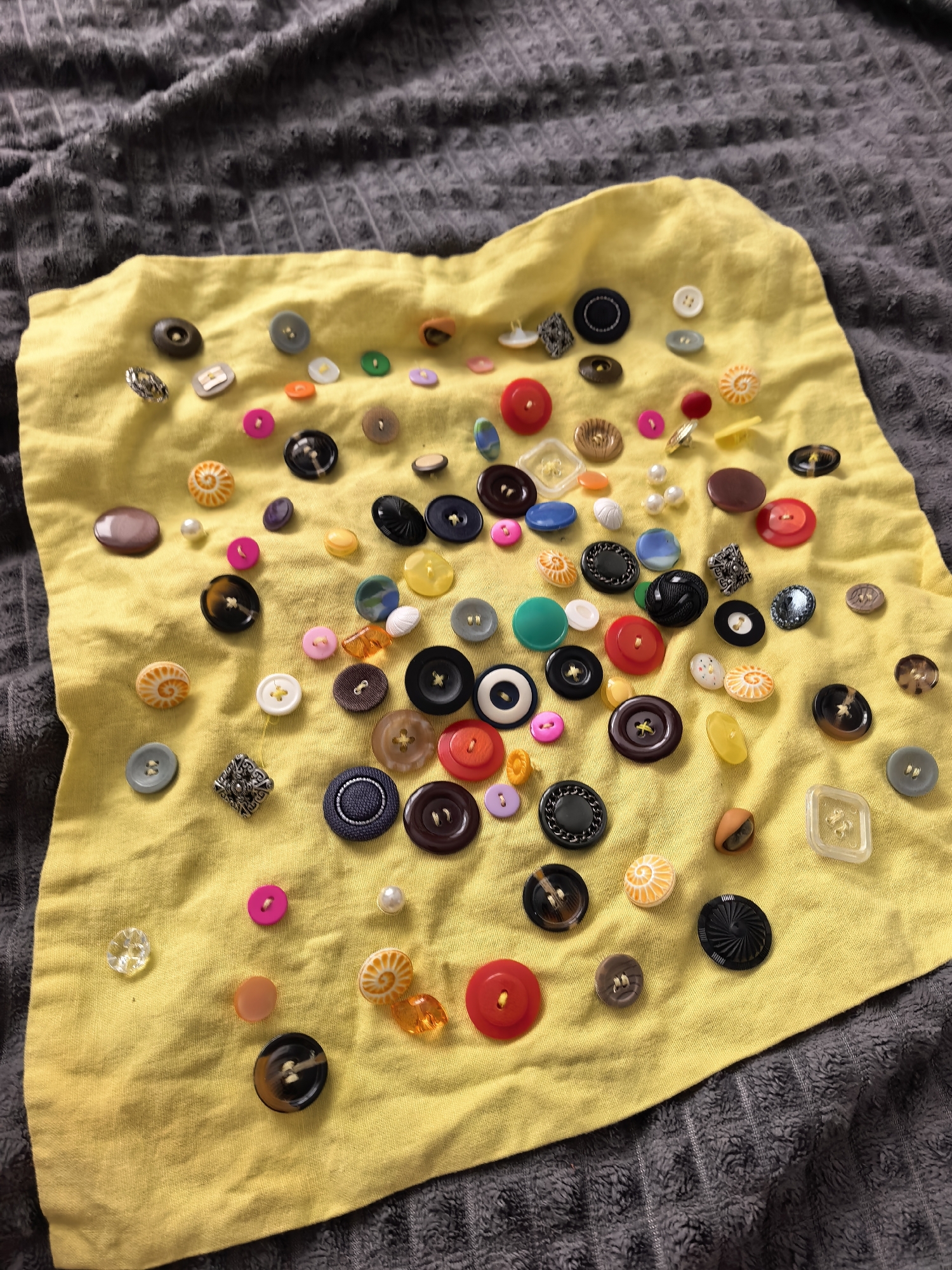 A top-down shot shows a yellow cloth resting on a gray textured blanket. The cloth is covered with a variety of buttons, differing in color, size, shape, and material. The buttons are scattered randomly across the cloth's surface. There are no people visible in the image. The image was taken in an unknown location and there is no information to suggest it was taken in Sterlitamak, Russia.
