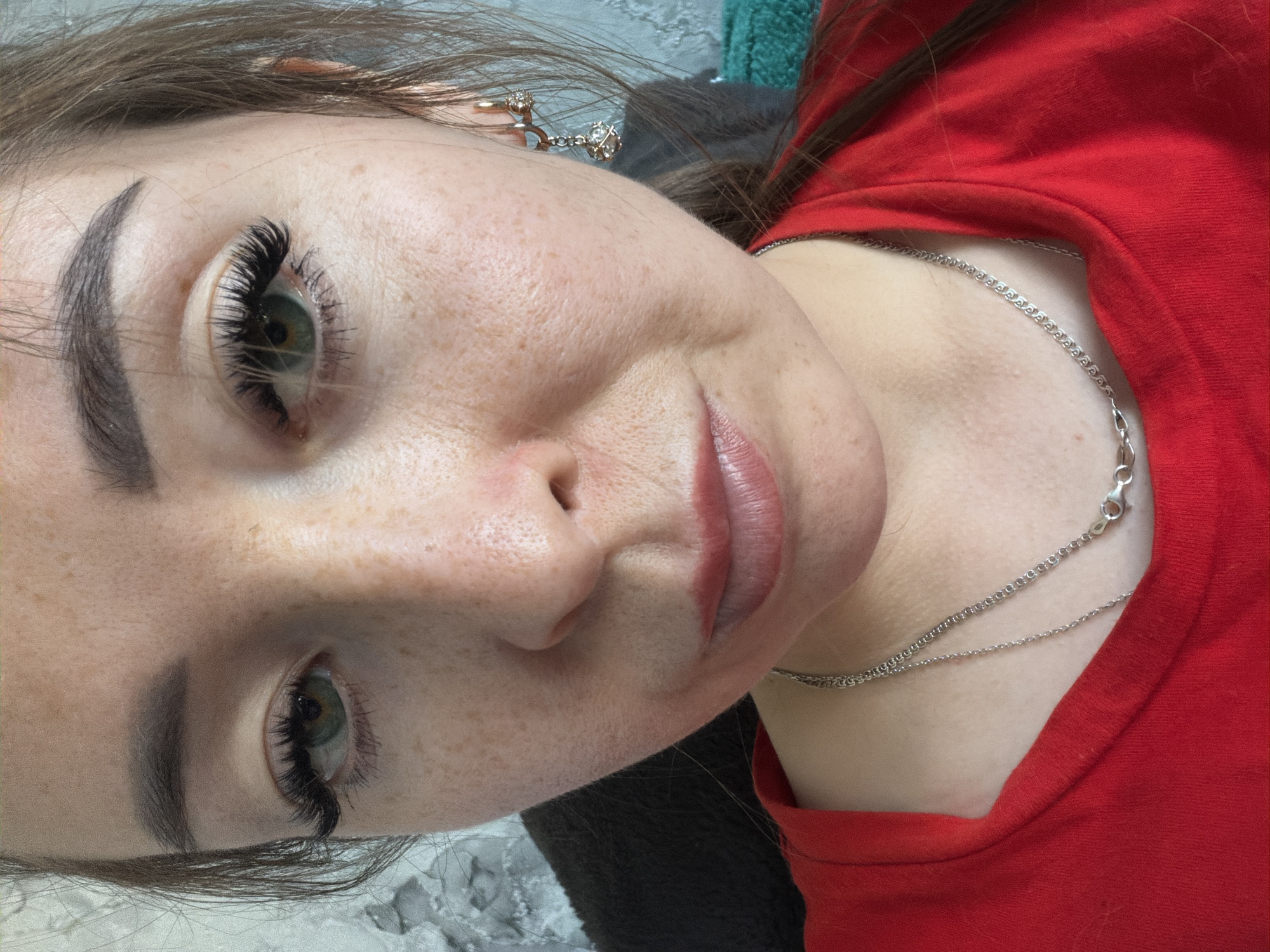 A medium close-up portrait shows a woman from the upper chest to the forehead. She is wearing a red t-shirt and a silver chain necklace. Her hair is brown and pulled back from her face. She has green eyes and is wearing false eyelashes. Her skin is fair with freckles. She is wearing gold hoop earrings with a diamond charm. The background is out of focus and appears to be a textured wall. There is no indication of the location in the city of Япрыково, Russia.
